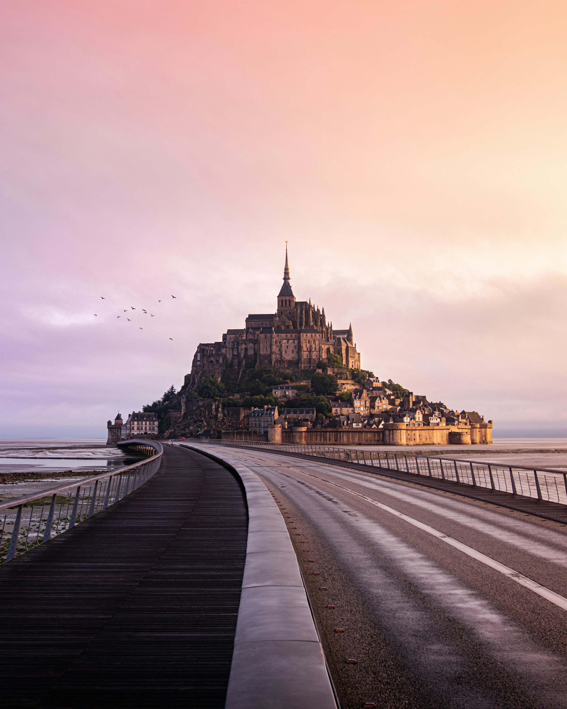 Vy över Mont Saint-Michel i Frankrike vid solnedgång med dramatisk himmel och tom väg som leder fram till ön.