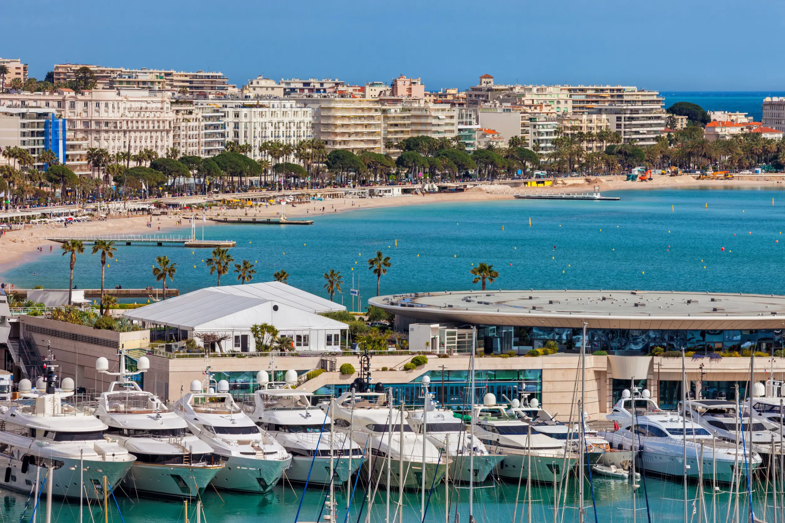 Resa till Cannes - Vy över en livlig marina med lyxbåtar förtöjda framför en sandstrand och en stadssilhuett med palmer och hotell i bakgrunden under en klarblå himmel.