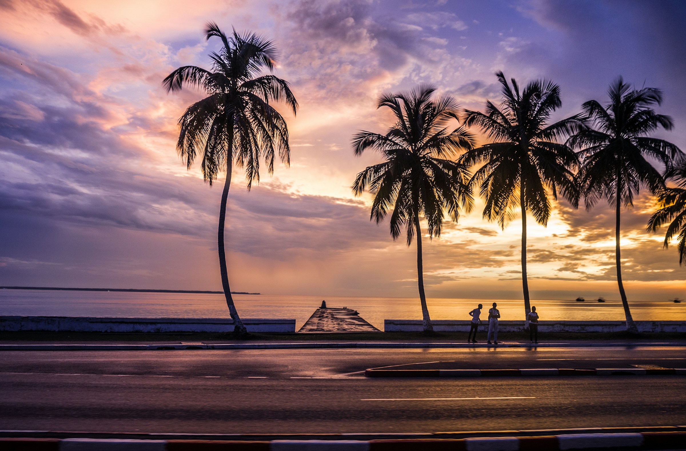 Solnedgång med siluett av palmer och gående personer vid strandpromenaden i Gabon.