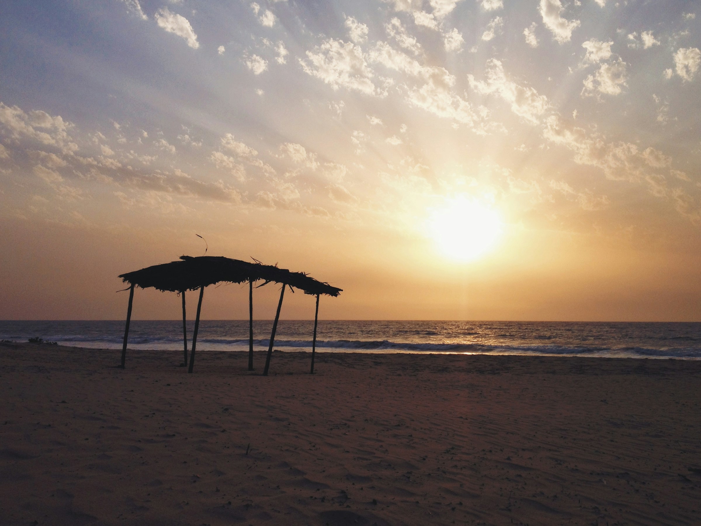 Strandparasoll av trä med halmtak mot en solnedgång på en sandstrand i Gambia