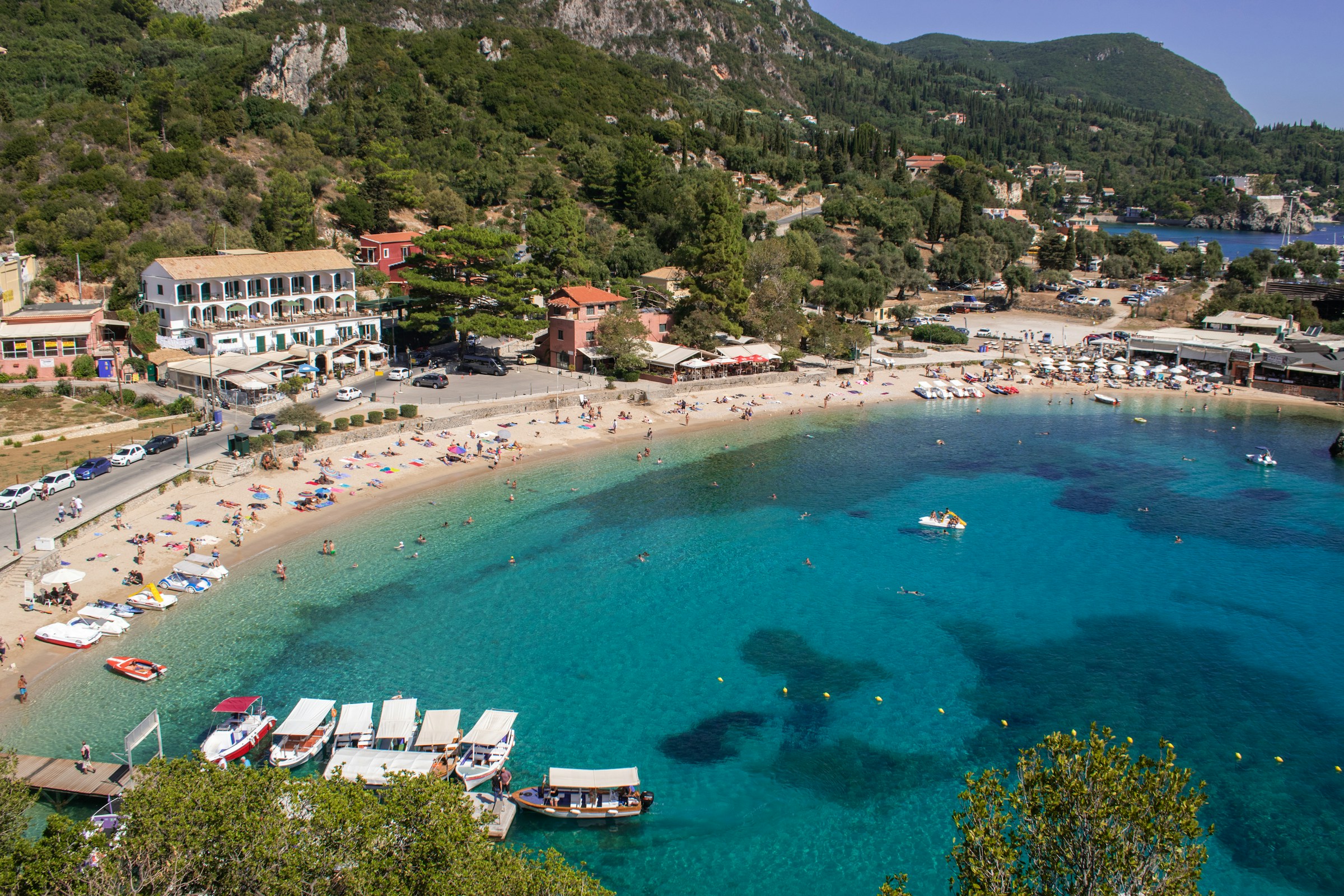 Livlig strand med kristallklart blått vatten, badgäster och båtar vid en pittoresk kuststad omgiven av gröna kullar på Korfu, Grekland