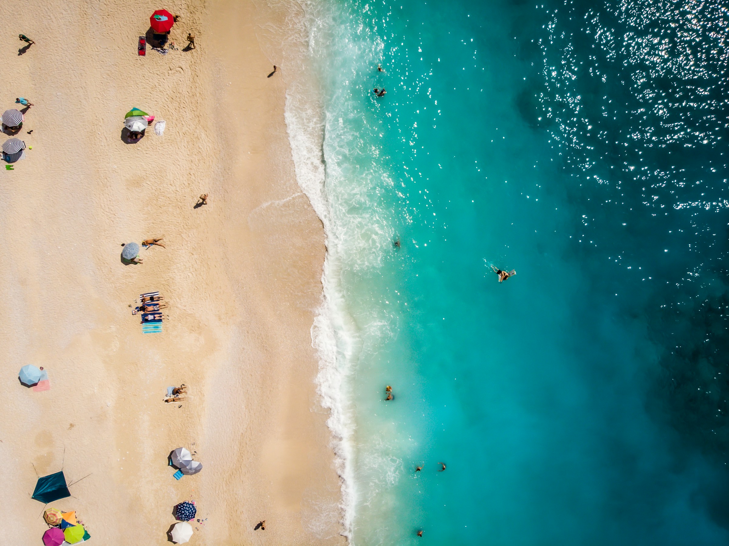 Vy över en livlig strand i Grekland med klart turkos vatten och människor som njuter av solen, fotograferad från ovan.