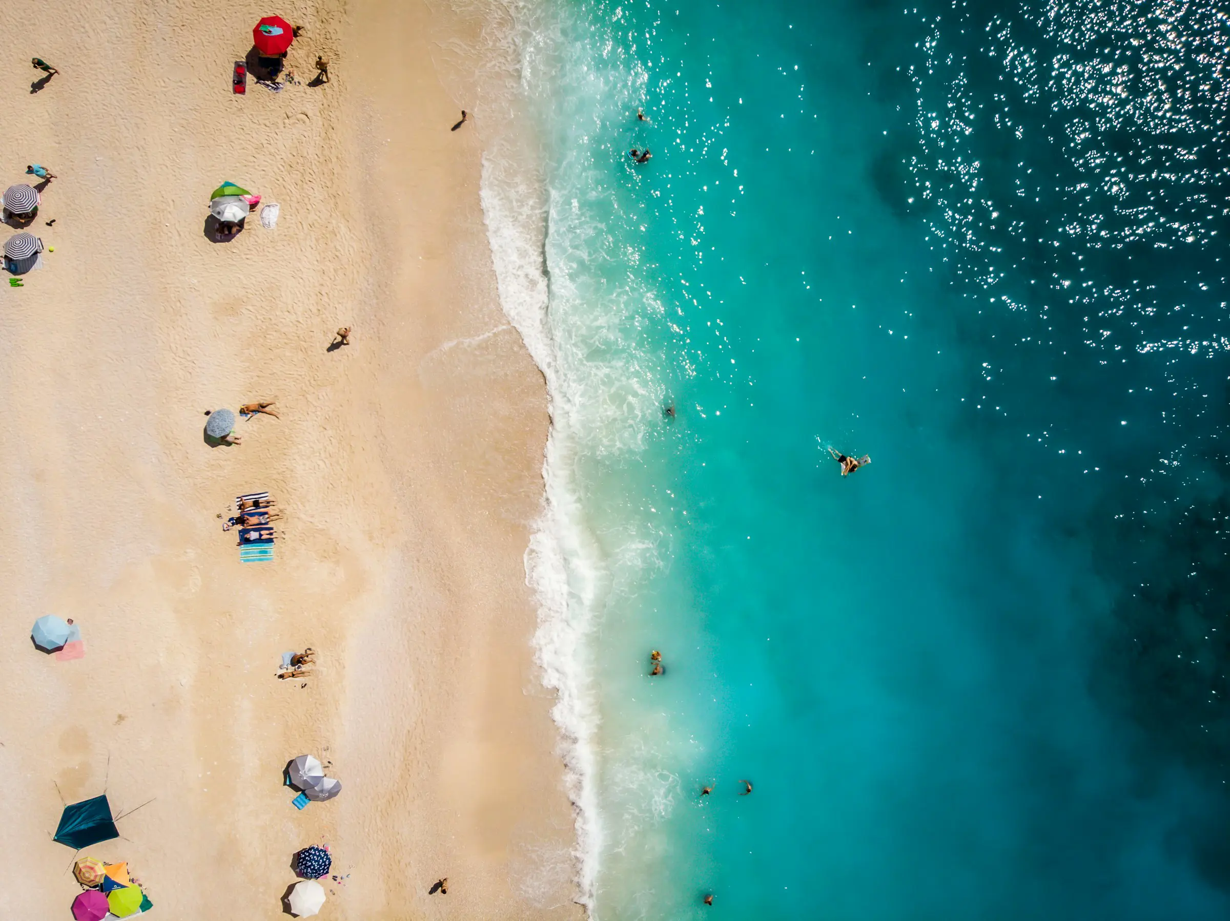 Aerial view of a beach with turquoise ocean water and sandy shore, dotted with sunbathers and colorful umbrellas.