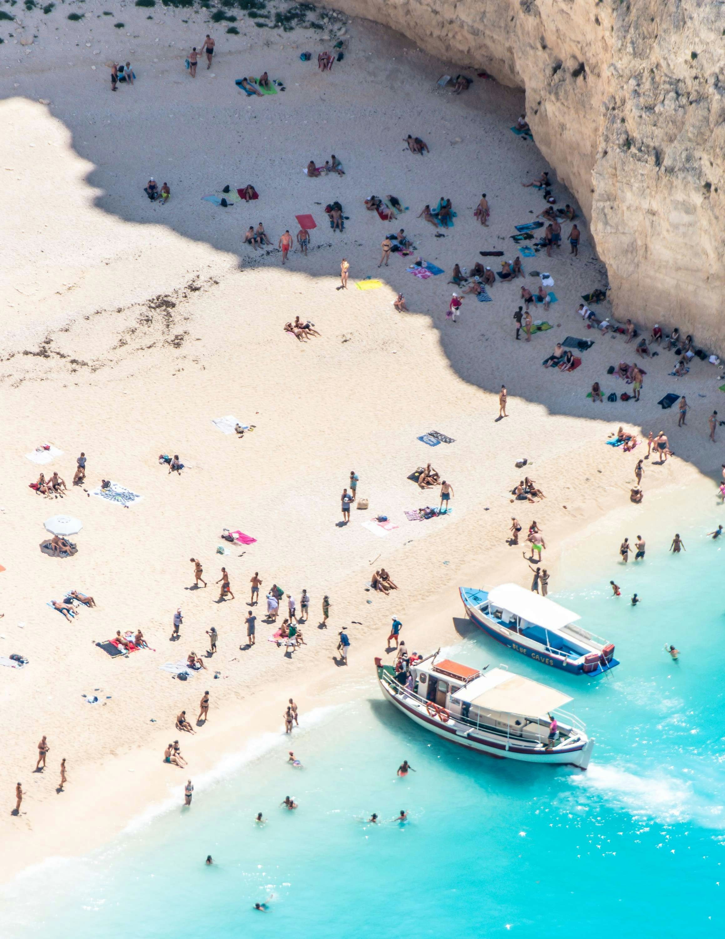 Flygbild av en livlig strand med klart turkosblått vatten och två fritidsbåtar nära kusten, full av badgäster och solbadare på en solig dag i Parga, Grekland