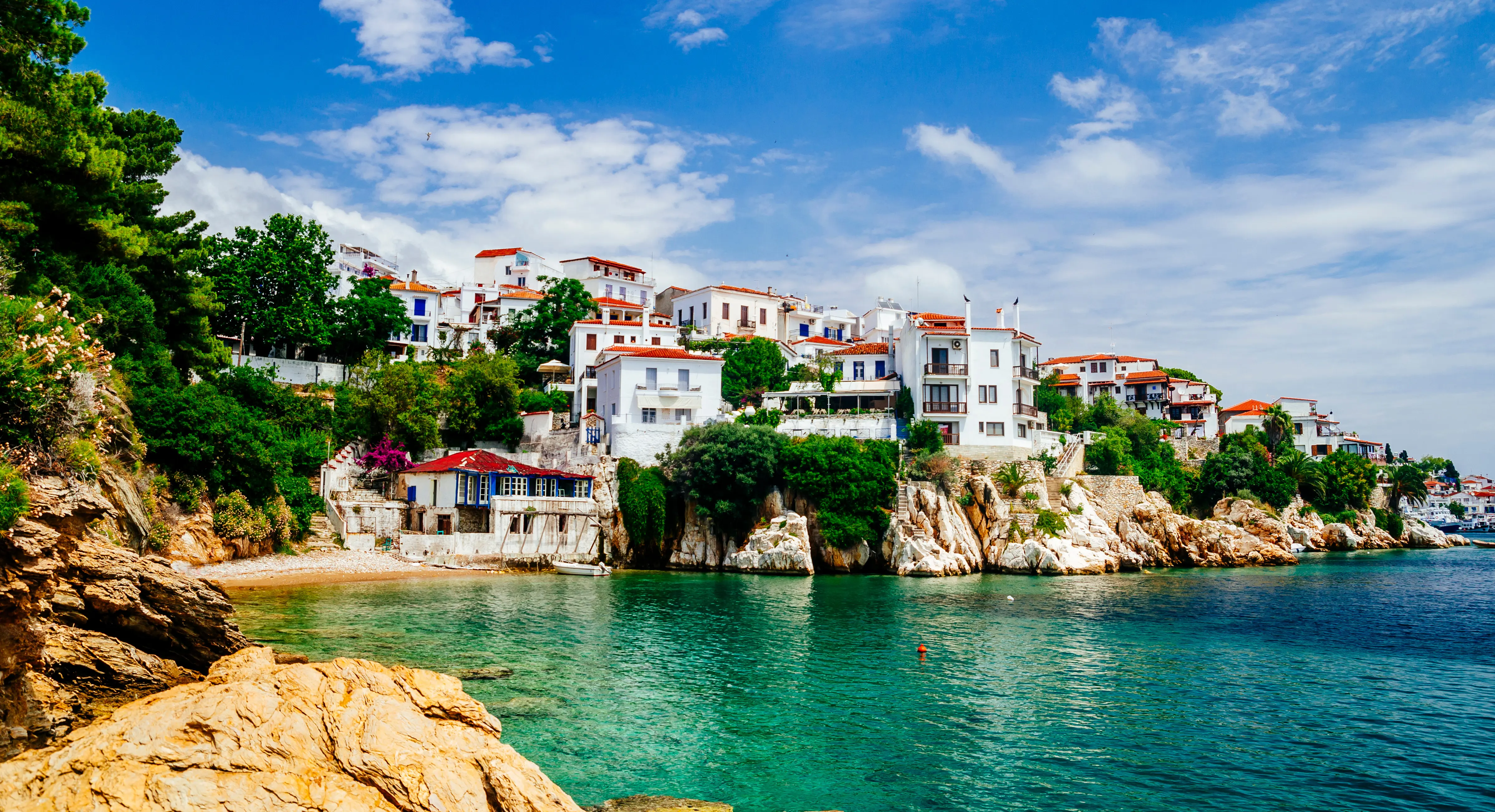 Resa till Skiathos - kuststad med pittoreska vitmålade hus vid klart turkosblått vatten, kantad av gröna träd och klippor under en blå himmel med lätta moln.