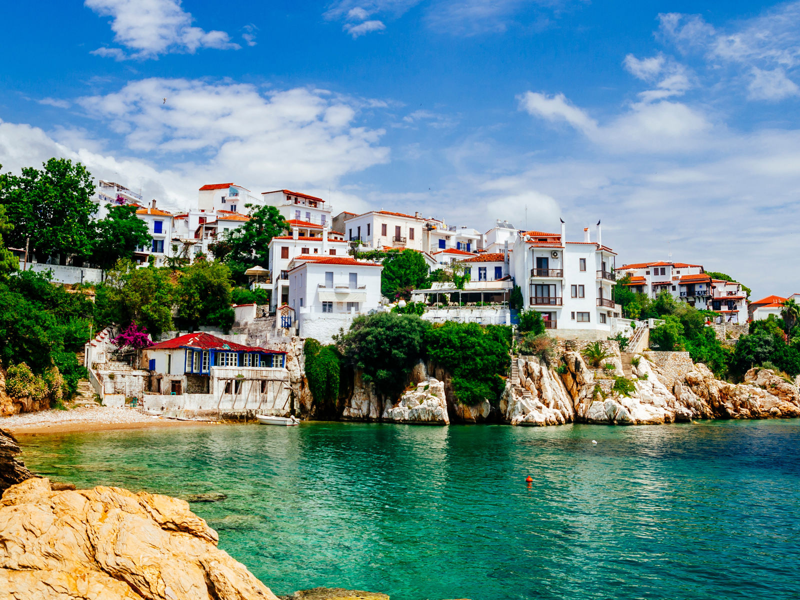 Resa till Skiathos - kuststad med pittoreska vitmålade hus vid klart turkosblått vatten, kantad av gröna träd och klippor under en blå himmel med lätta moln.
