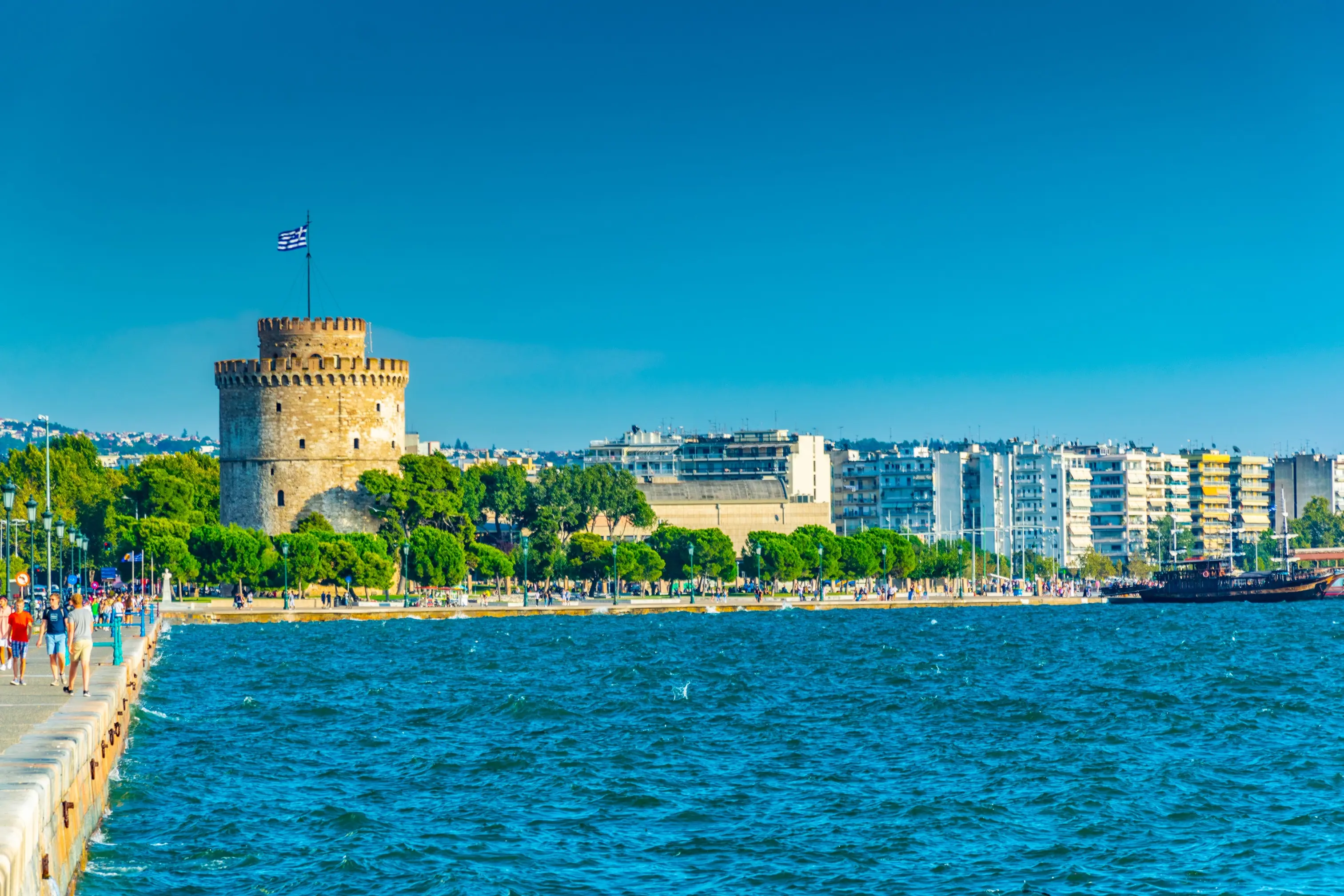 Vy över den vitkalkade tornet Vita Tornet i Thessaloniki, Grekland, med promenerande människor längs kustpromenaden och stadens arkitektur i bakgrunden under en klarblå himmel.