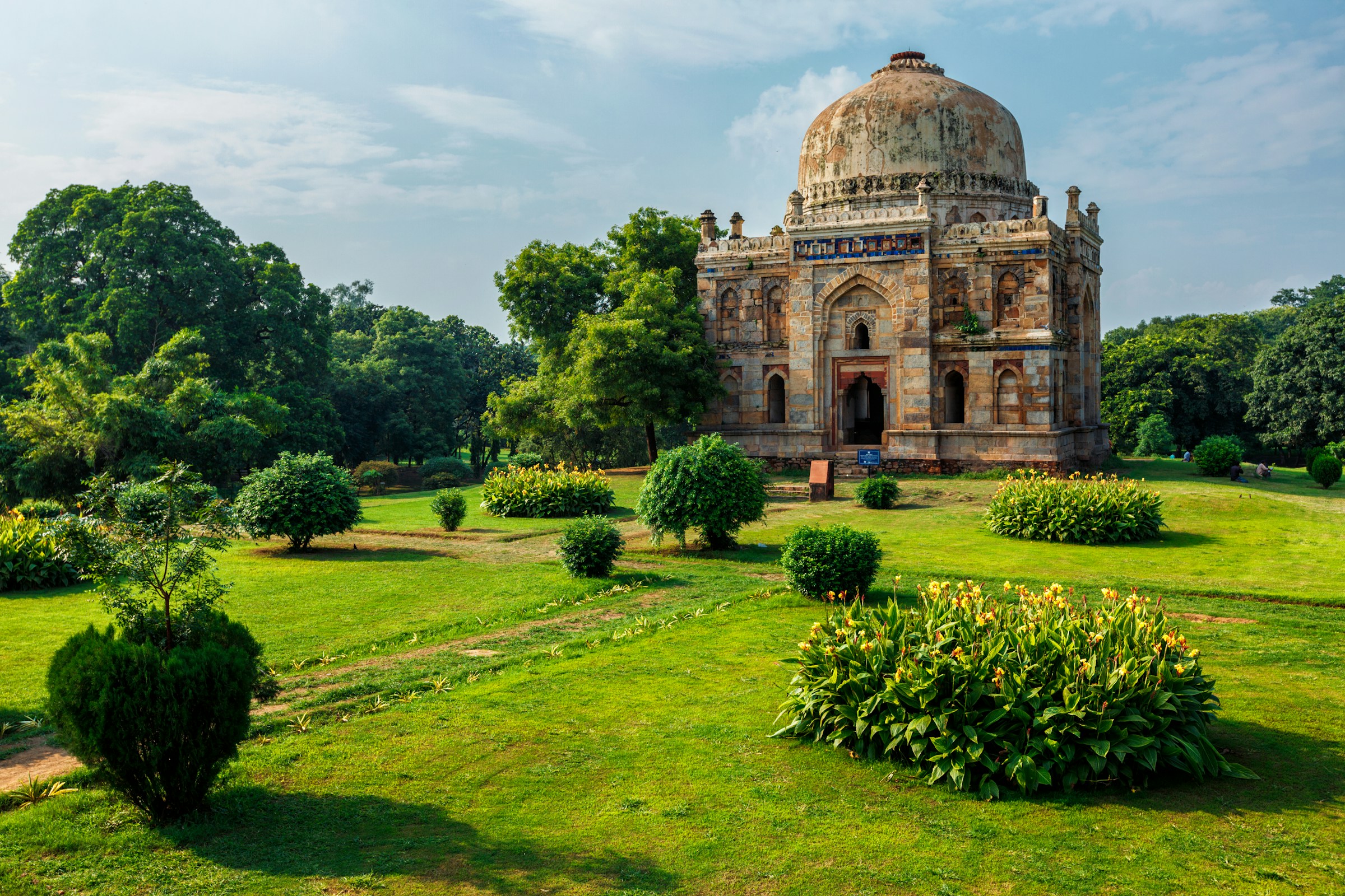 Gammalt historiskt mausoleum omgivet av gröna trädgårdar och växtlighet under solig himmel i Indien.