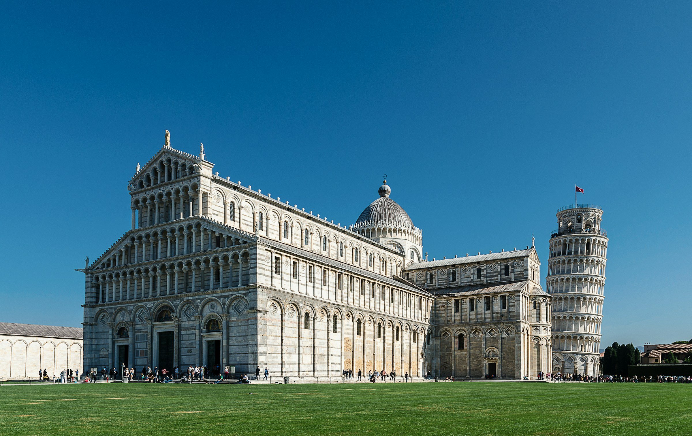 The famous Leaning Tower of Pisa stands on a green lawn with a kyra next to it against a blue sky