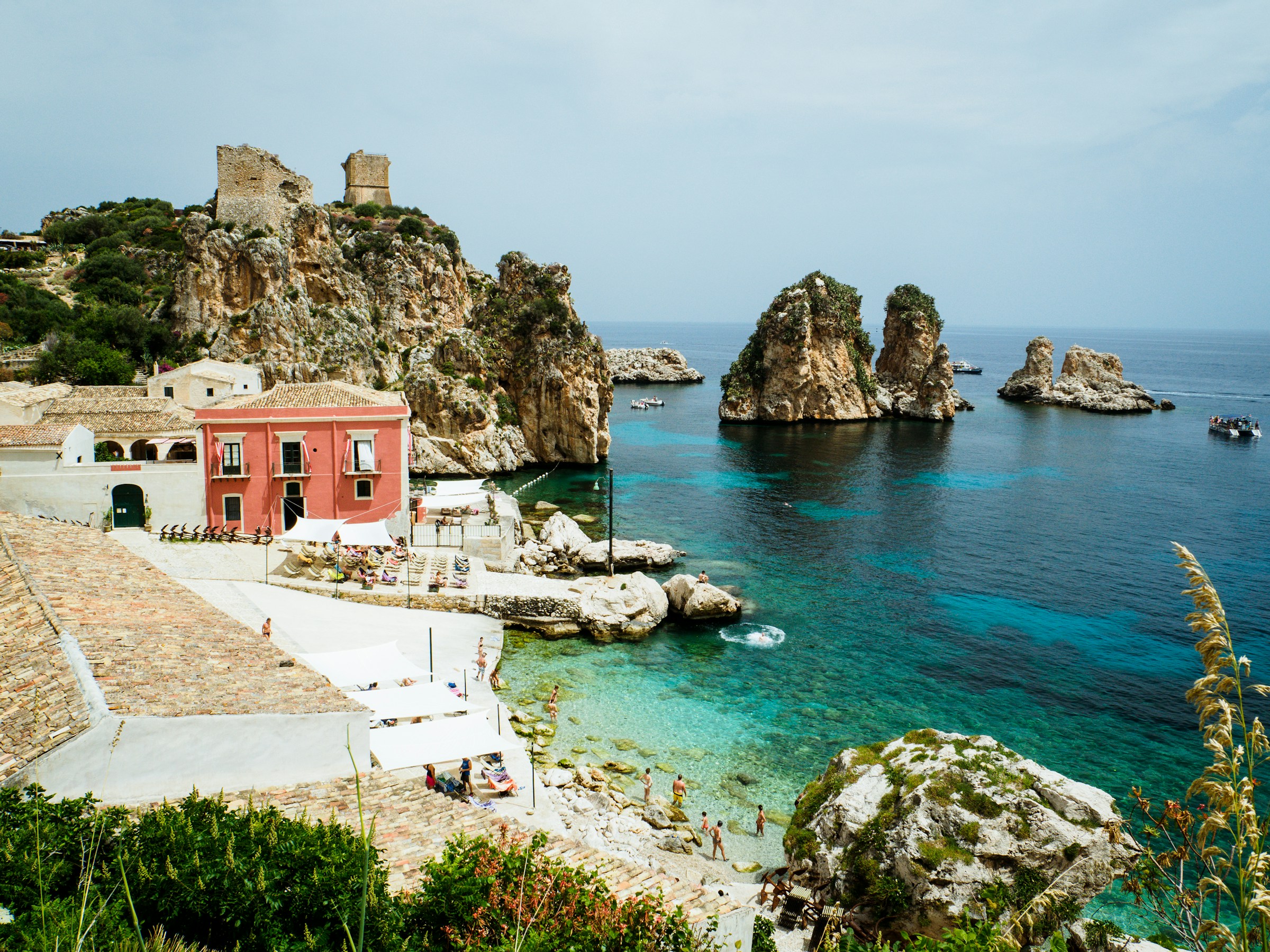 Idyllisk medelhavsutsikt med klippor, ett gammalt vakttorn, kristallklart vatten och en röd byggnad vid kusten av Sicilien.