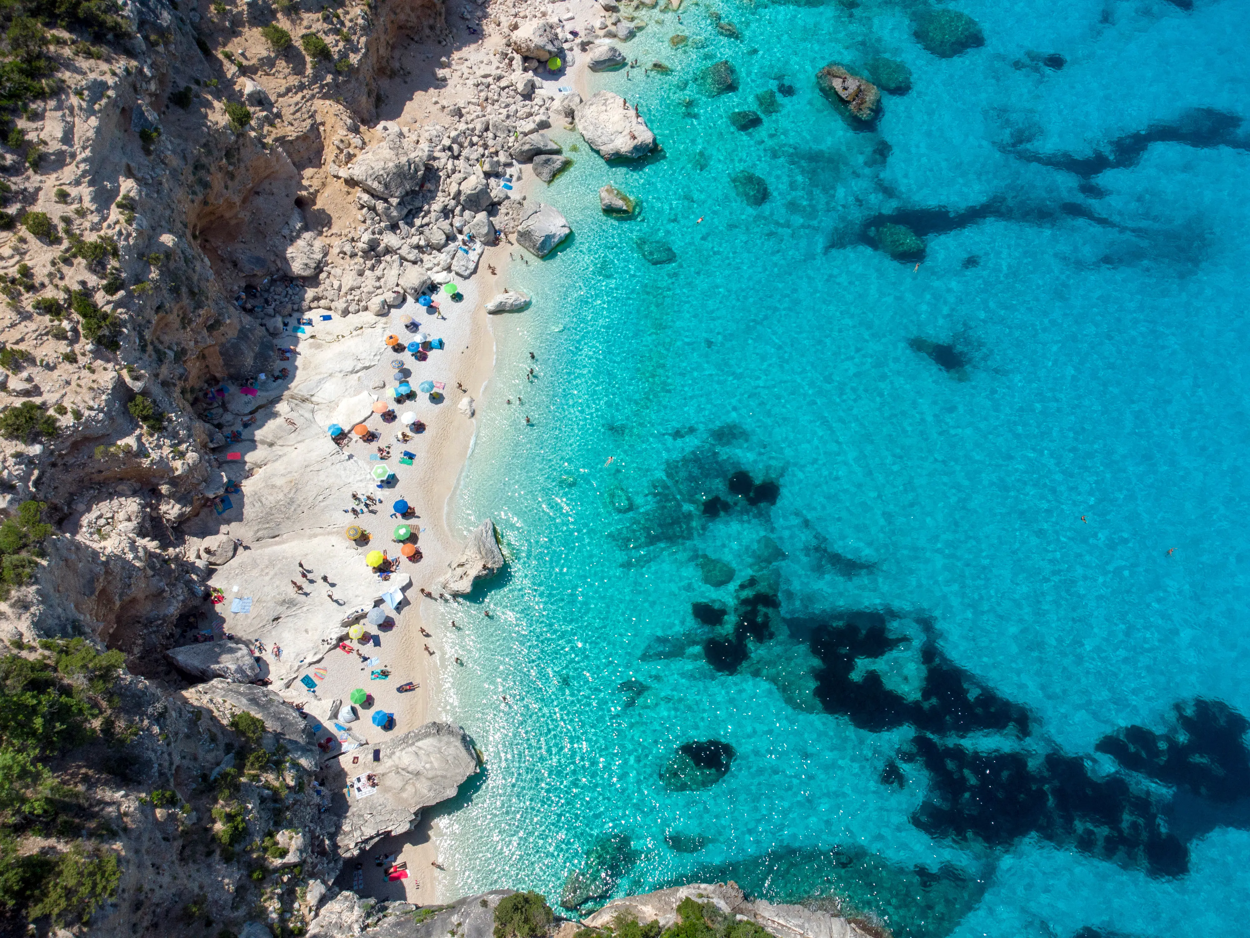 Resa till Sardinien - Flygfoto av en klarblå lagun och strand med badgäster och parasoll.