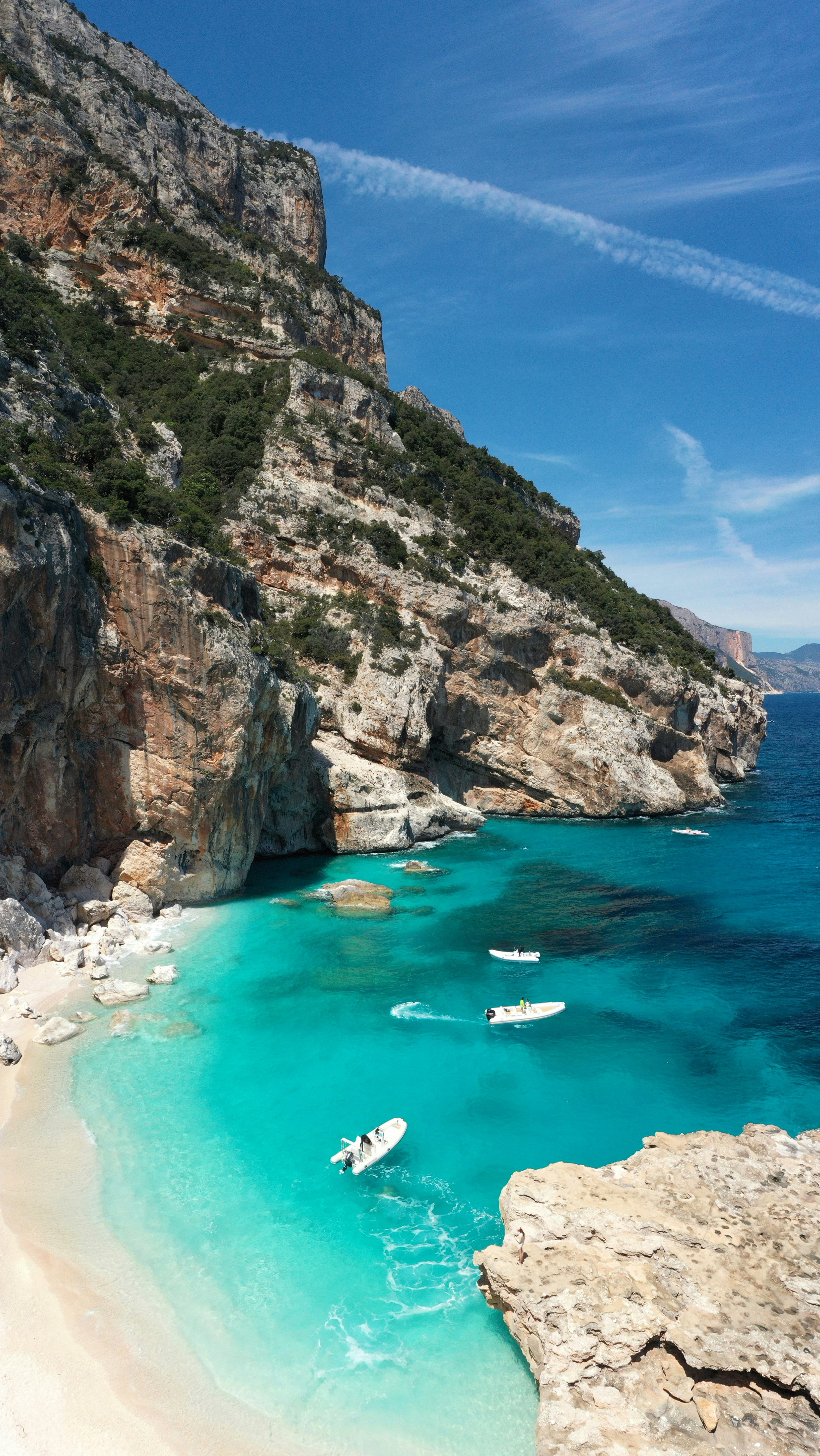 Turkost vatten omger klippiga kustlinjen med små båtar som flyter nära stranden på en solig dag på Sardinien.