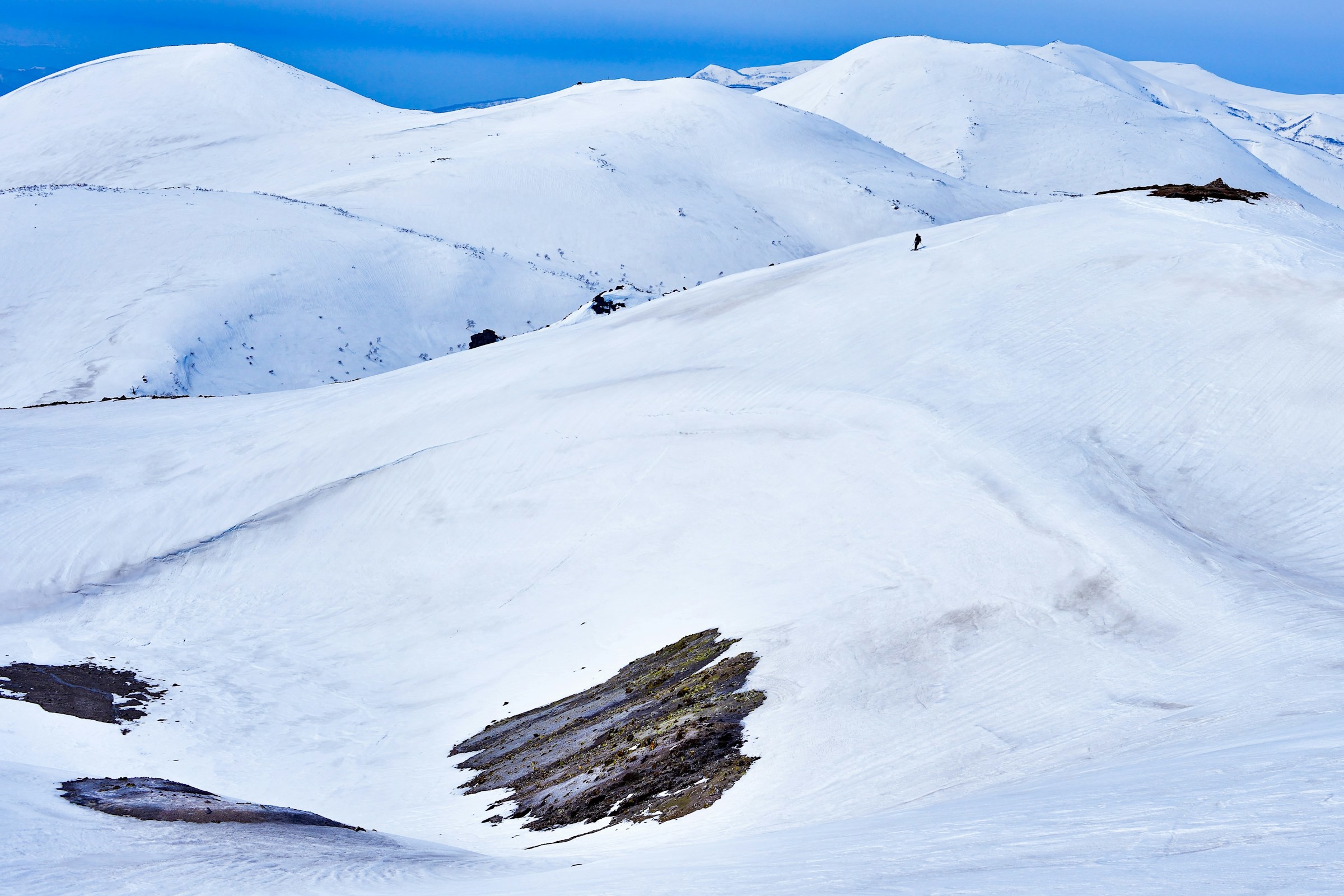 Snow-capped mountain peaks under a clear blue sky, with traces of rock formations and a lone hiker in Niseko, Japan.