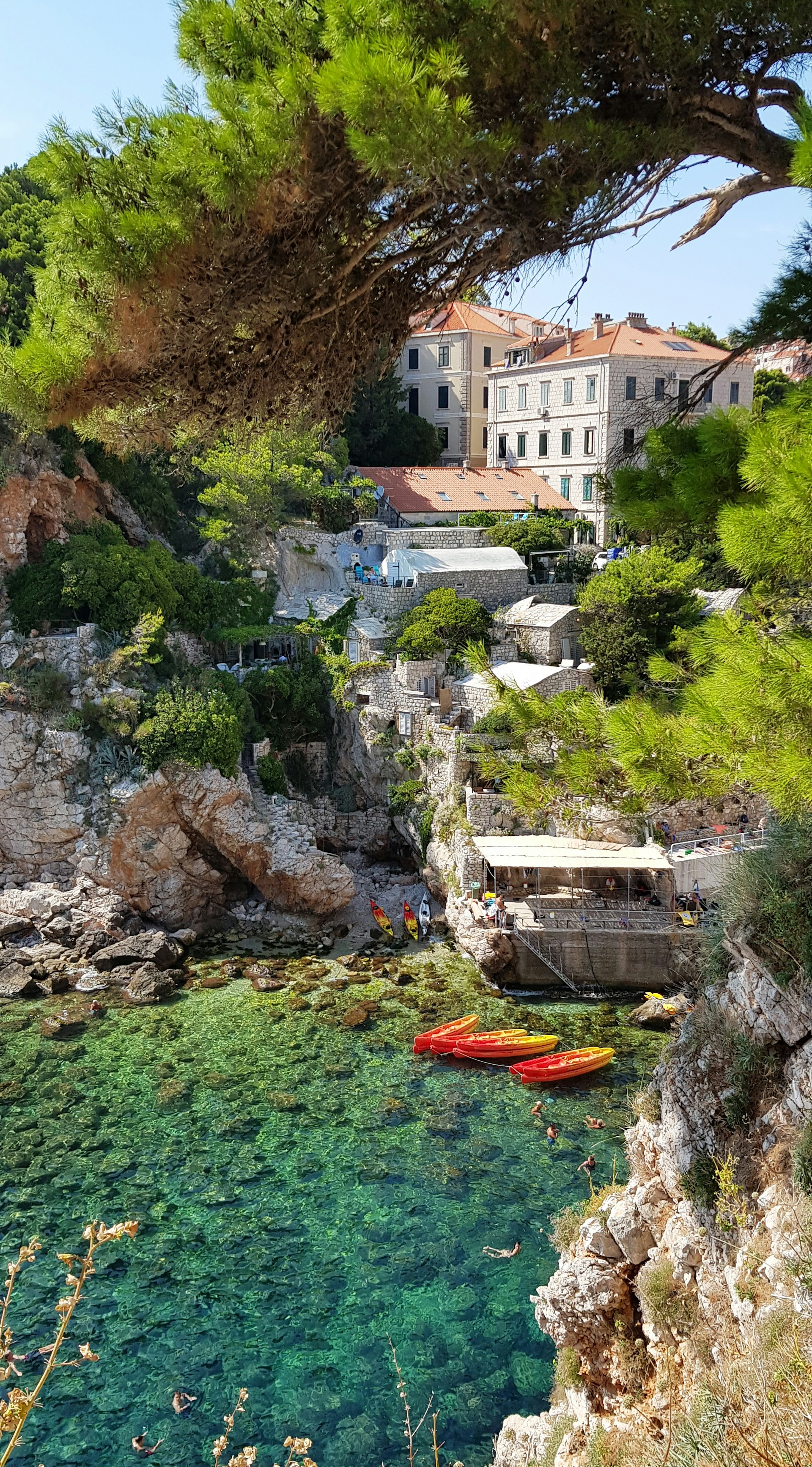 Klarblått hav vid en klippig bukt med gröna träd, röda kajaker och traditionella stenhus i Dubrovnik.