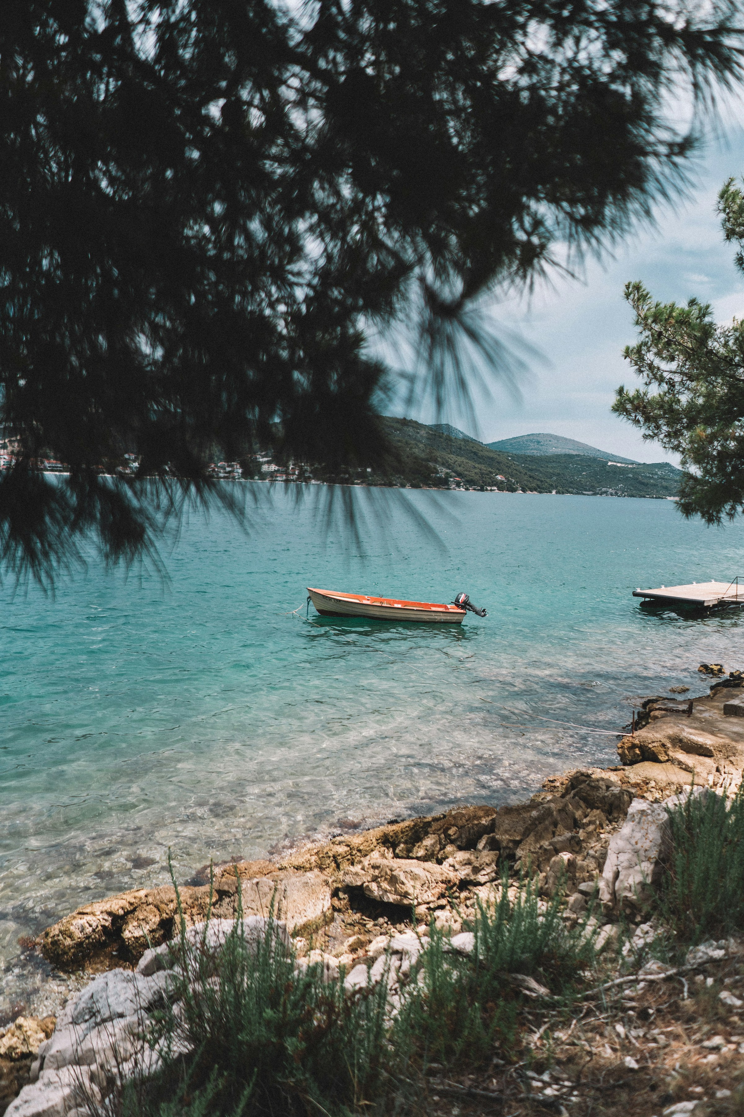 Ensam röd båt förtöjd i klart turkos vatten nära stenig kust med lummiga träd i förgrunden och kullig landskap i bakgrunden i Split.