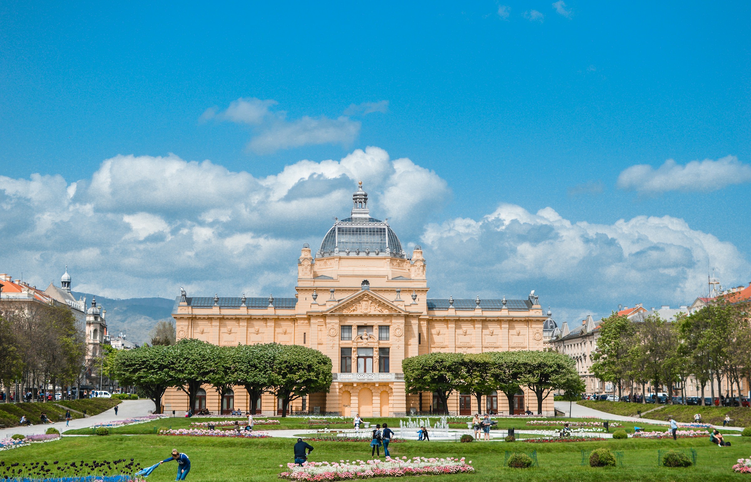 Elegant historisk byggnad med kupoltak mot en blå himmel omgiven av en välskött offentlig trädgård med blomrabatter och besökare i Zagreb.