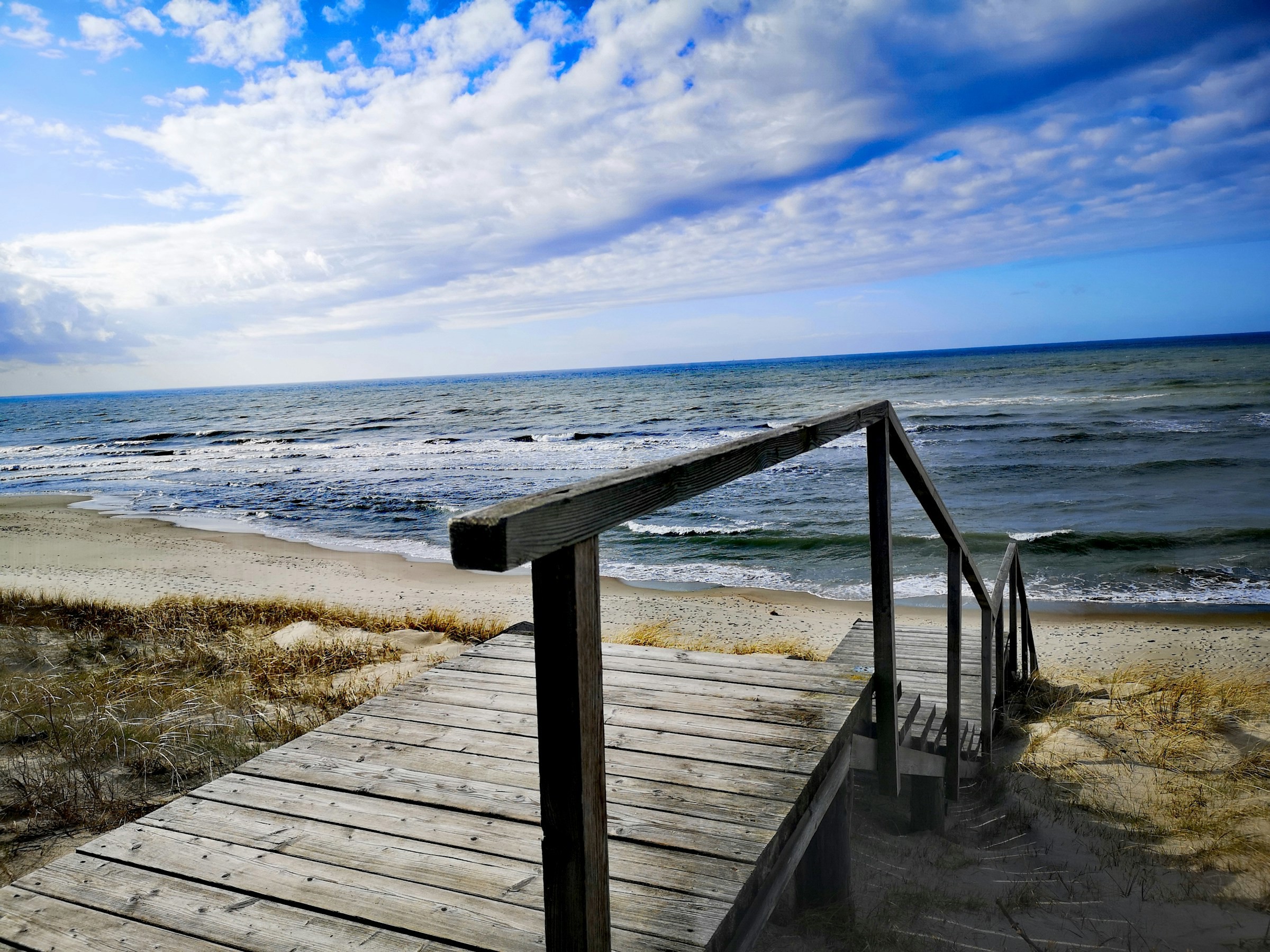 Trätrapp som leder till en sandig strand vid havet under en molnig himmel i Litauen.