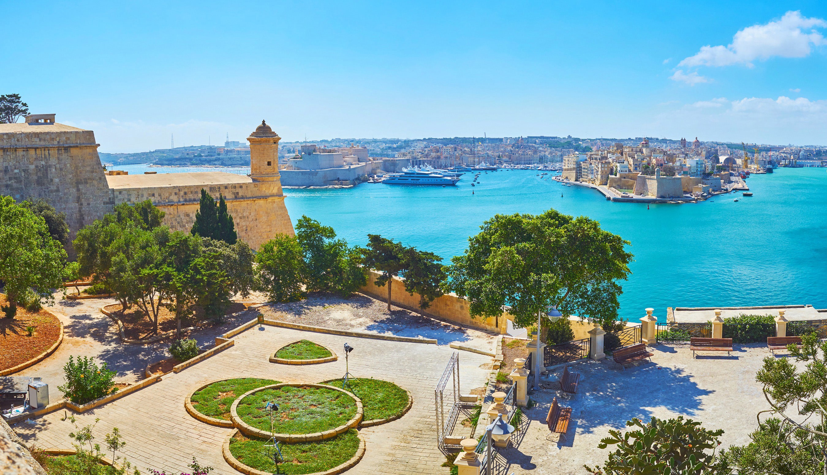 En vacker trädgård och utegård med grönska blickar ut över turkosblått hav vid kanten av en stad på Malta