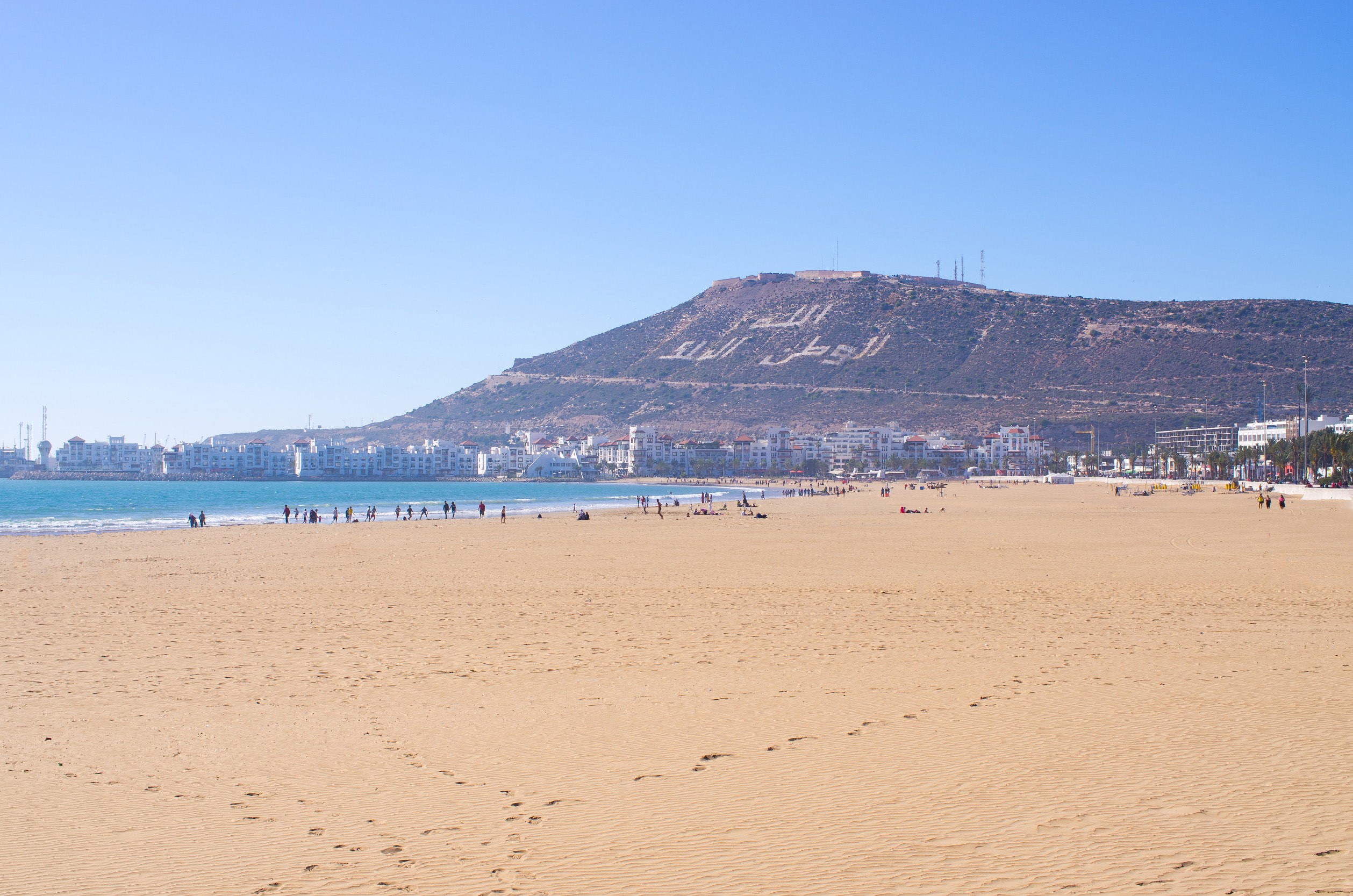 Stor sandstrand vid kusten av Agadir med blått hav och stora berg i bakgrunden vid horisonten