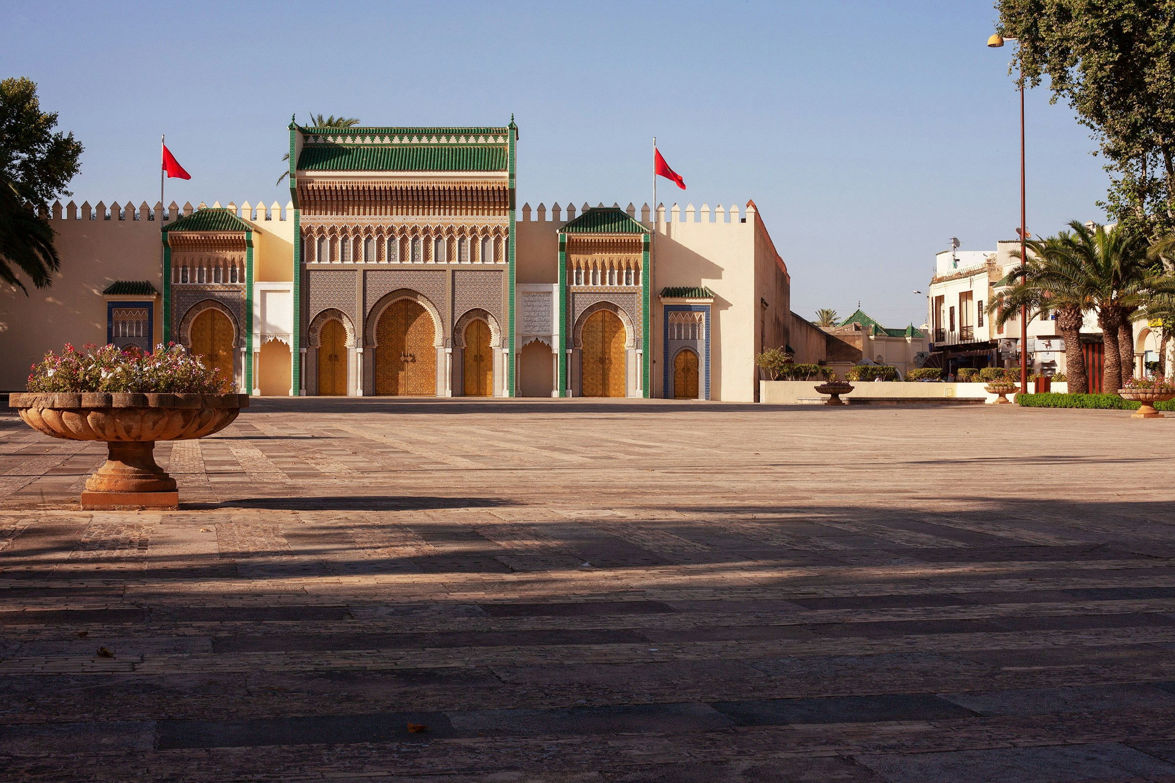 Vacker arkitektur på byggnad med gröna och vita mosaiker och Marockanska flaggor, sedd från en stenlagd gata med blomkruka i förgrunden.