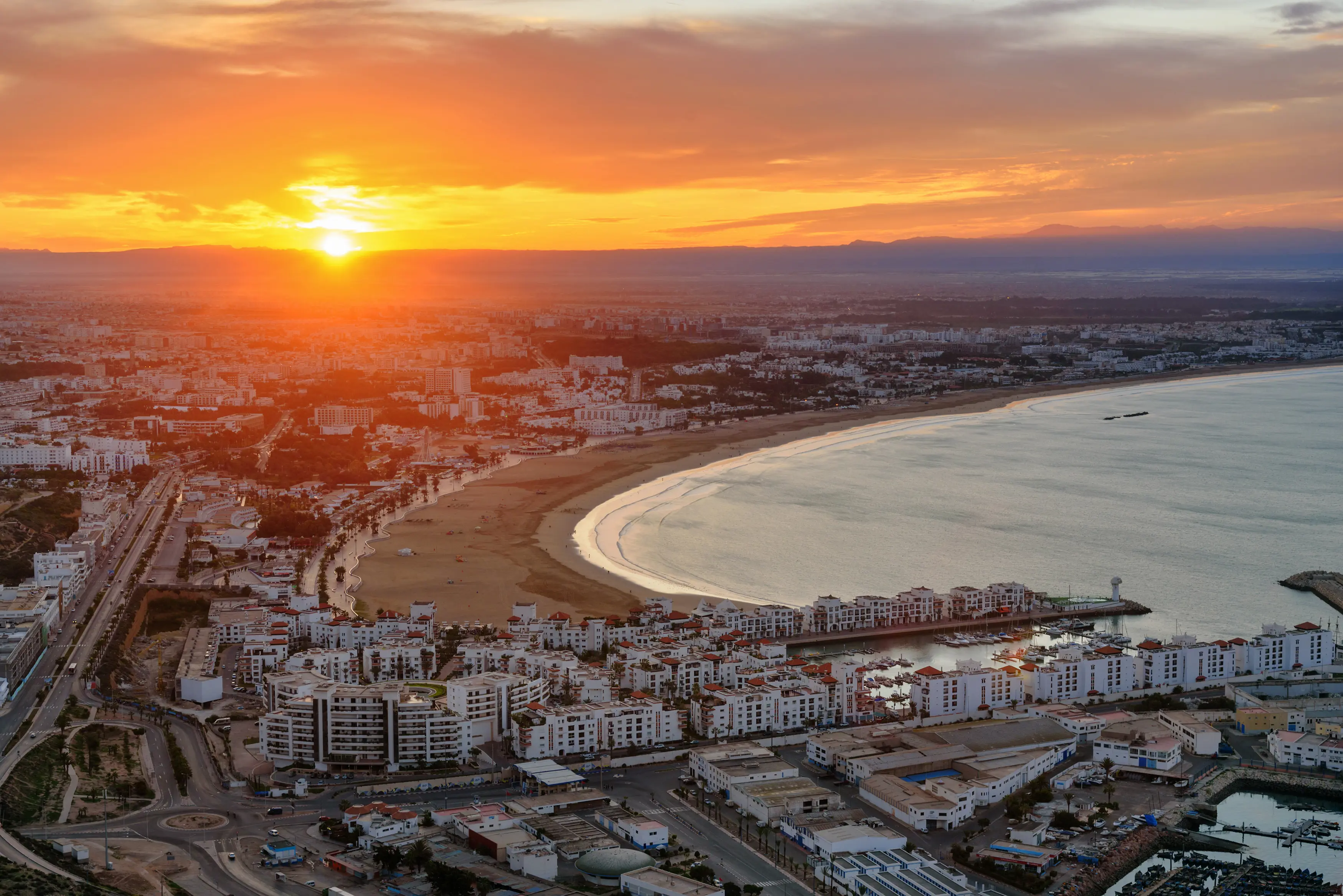 Resa till Agadir - Flygfoto av en kuststad vid solnedgång med dramatisk himmel och reflexer i havet, vita byggnader och en strandpromenad som sträcker sig längs kusten.