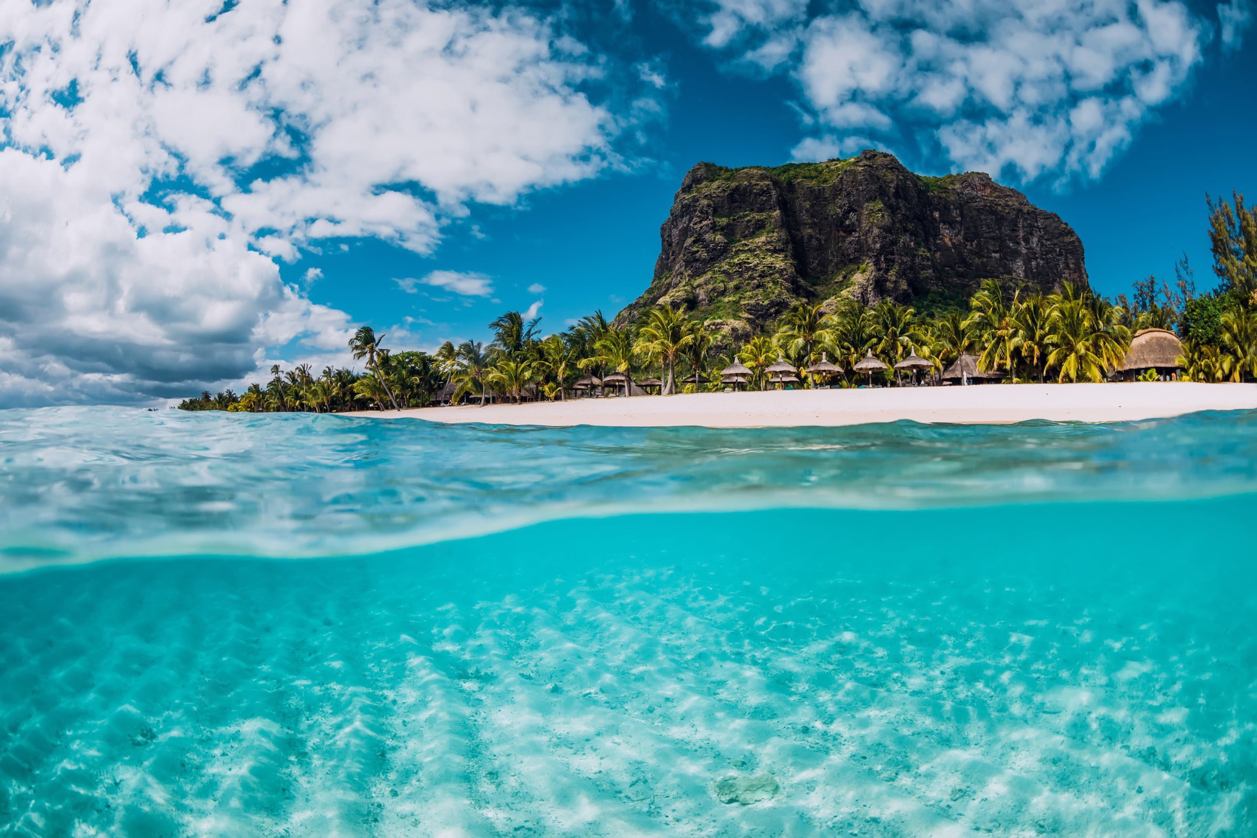 Vy från under turkosblått vatten upp mot en ö och strand på Mauritius med blå himmel i bakgrunden