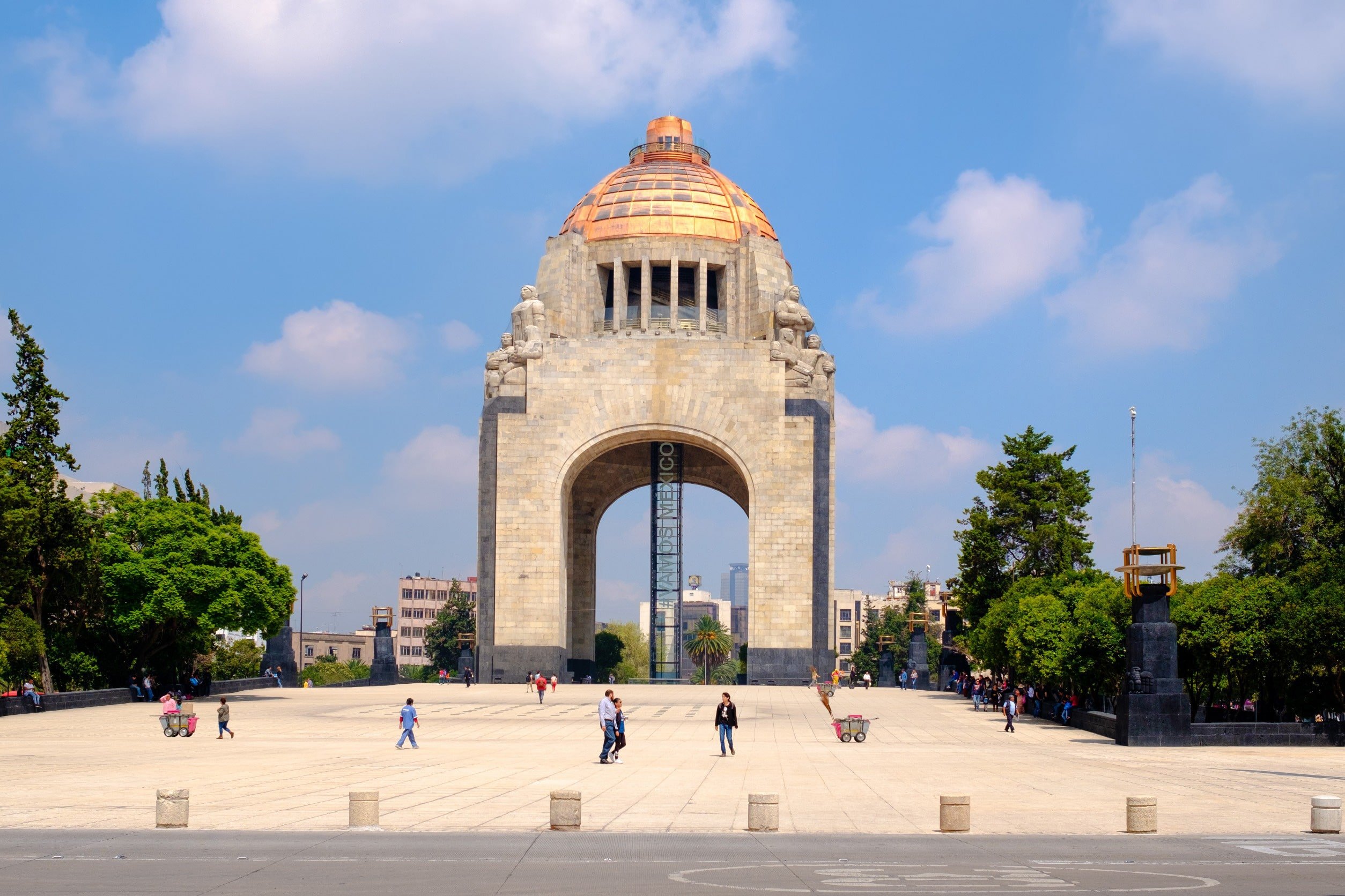 A square leading up to monuments in Mexico City with green trees around and blue sky in the background