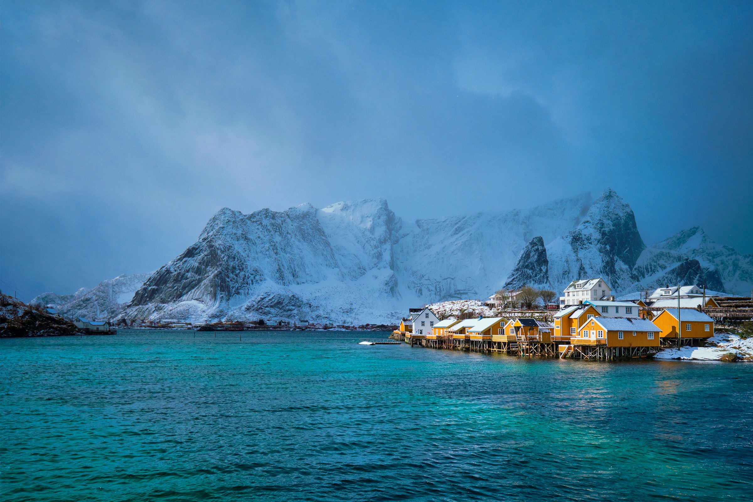 Vy från vattnet upp mot upplysta stugor med snötäckta berg i bakgrunden och en molnig himmel