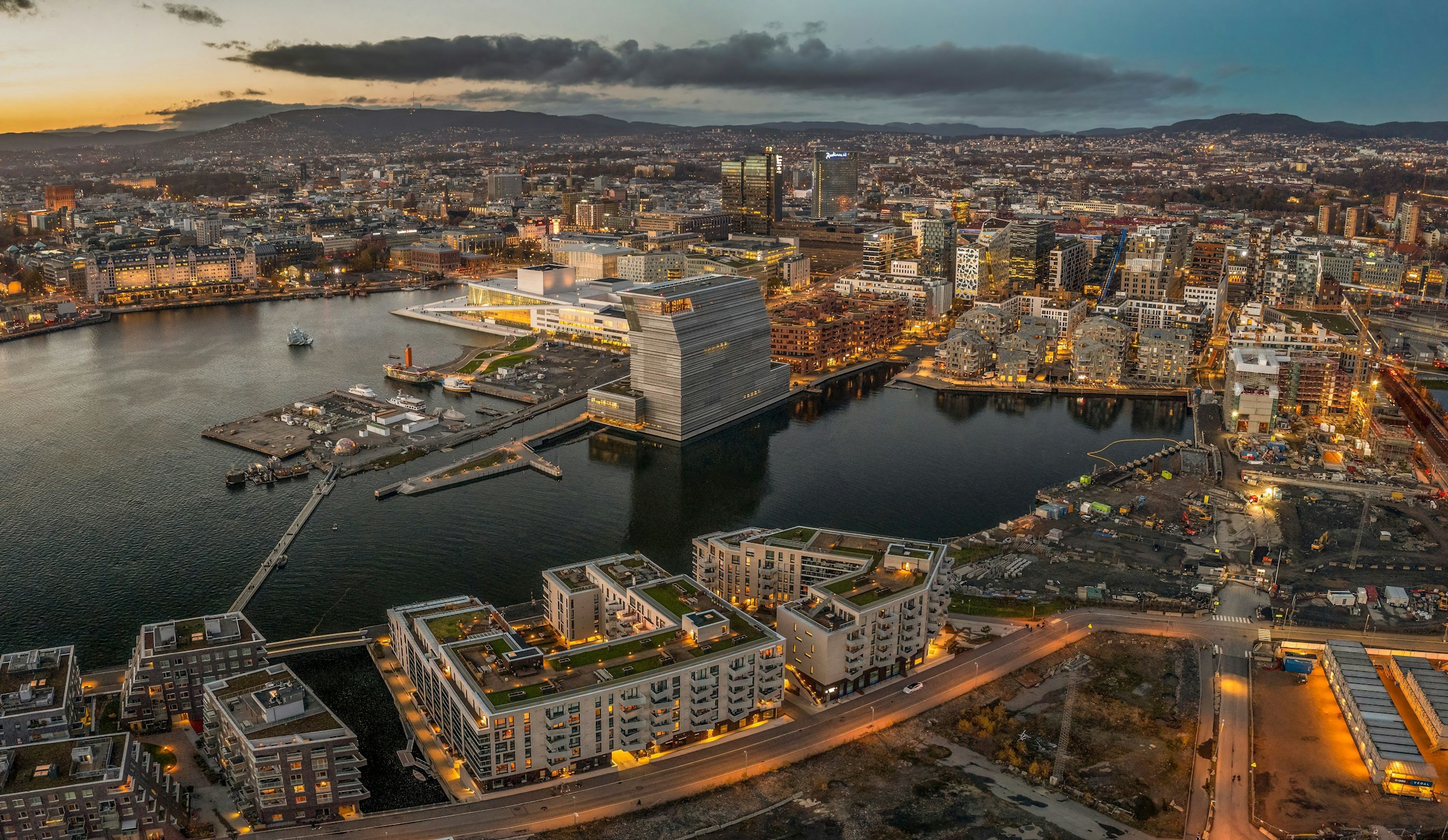 Fågelvy över Oslo-stad under en solnedgång med en hamn och vackert vatten och lyssande byggnader