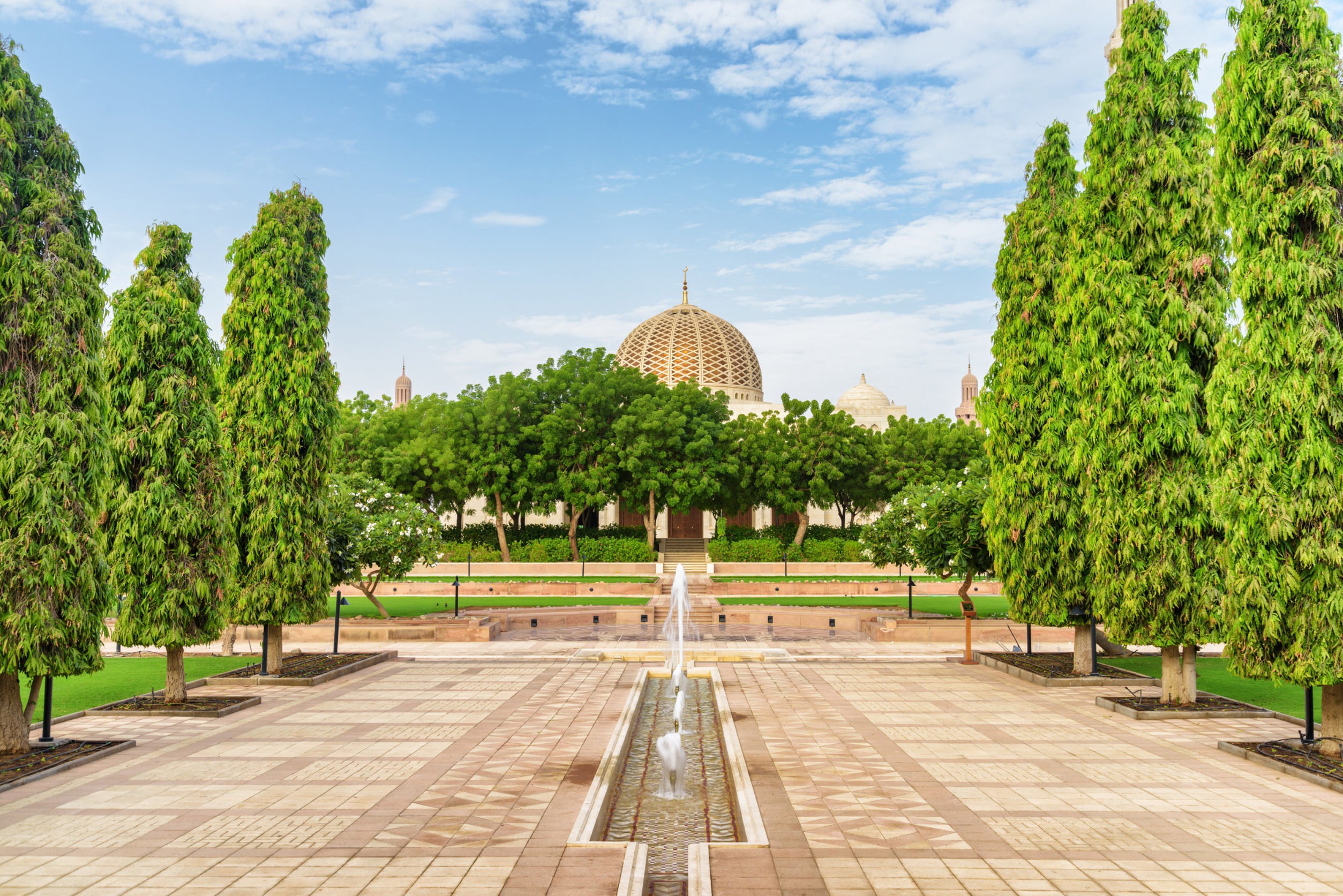 Wide street with green trees along the edges leads to a garden and a mosque in the background