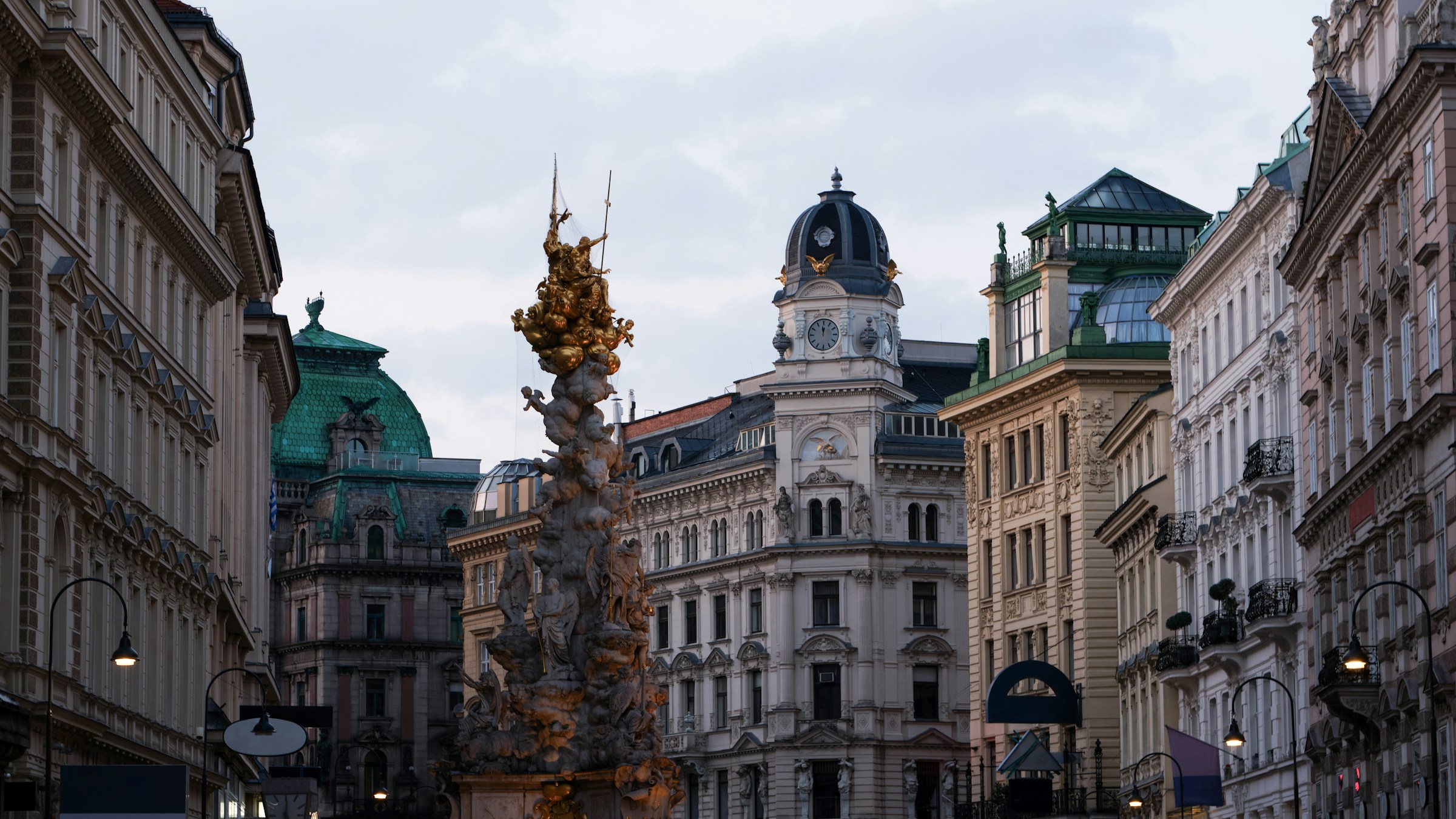 Vy över en gatustratning i Wien med en intrikat gyllene pestkolonn och klassiska byggnader med kupoler och fasader, vid skymning."