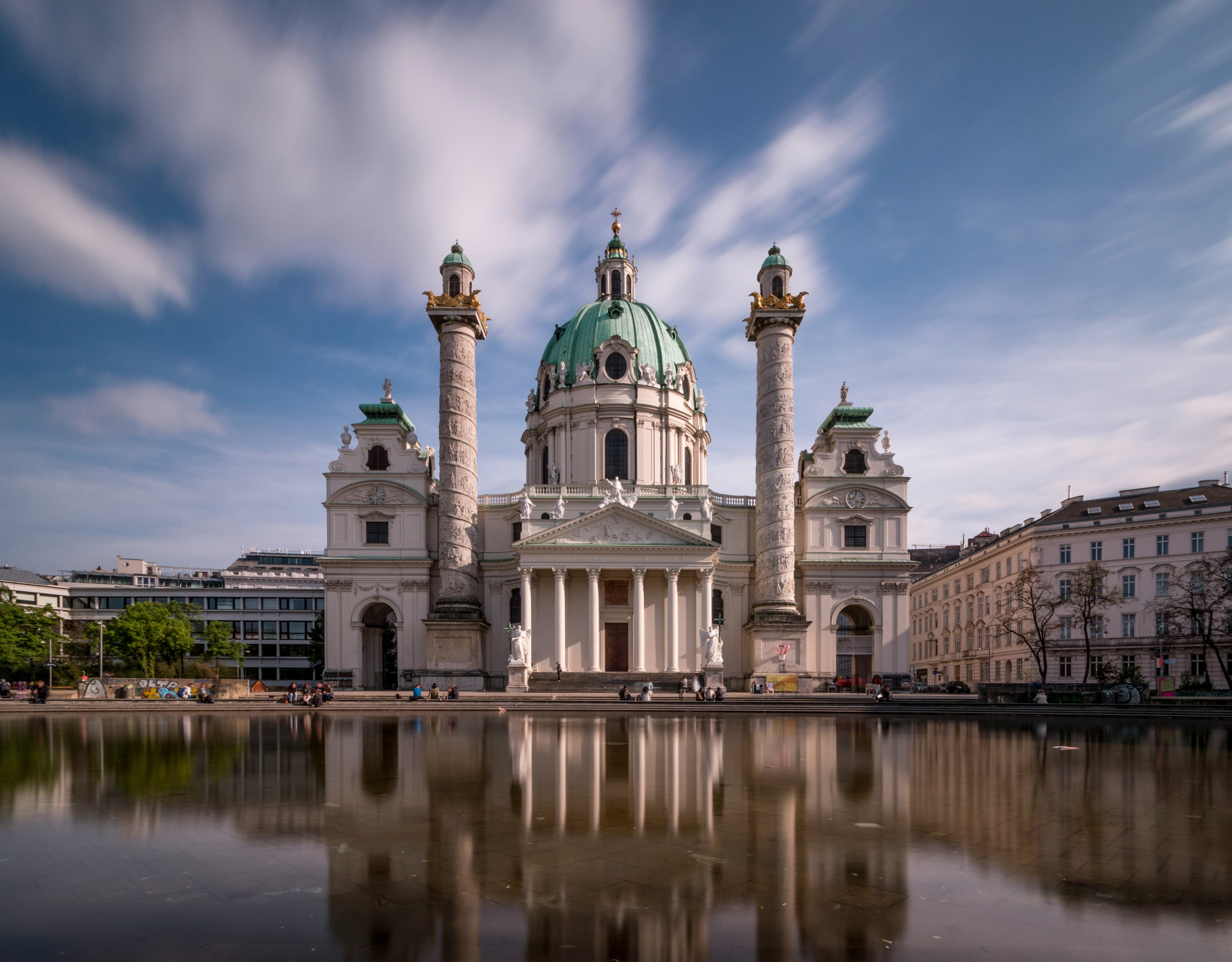 Karlskirche med reflektion i dammen på Karlsplatz i Wien, med moln i rörelse på en solig dag.