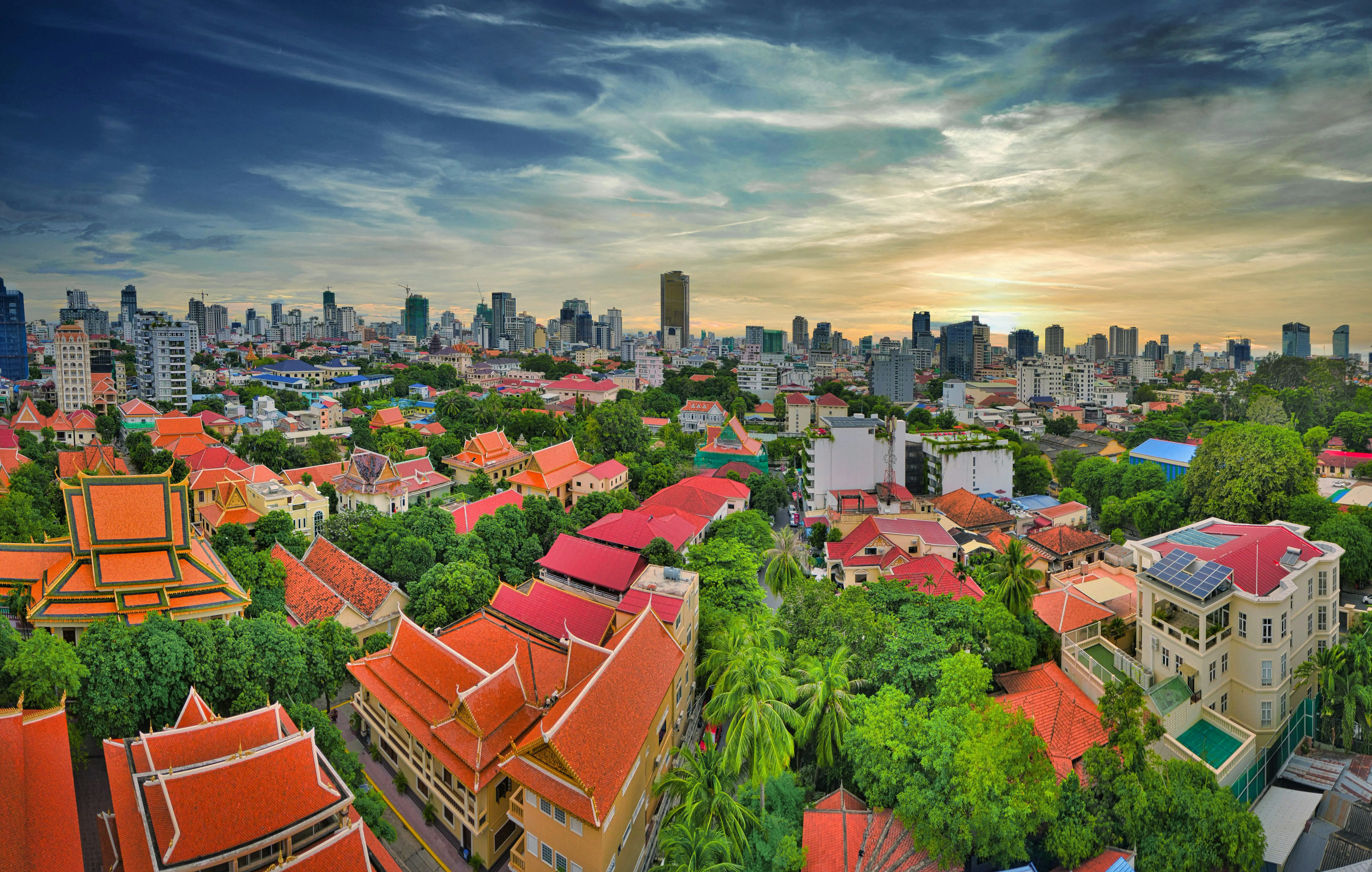 Travel to Phnom Penh - Panoramic views of Phnom Penh at sunset with traditional and modern buildings surrounded by lush trees.