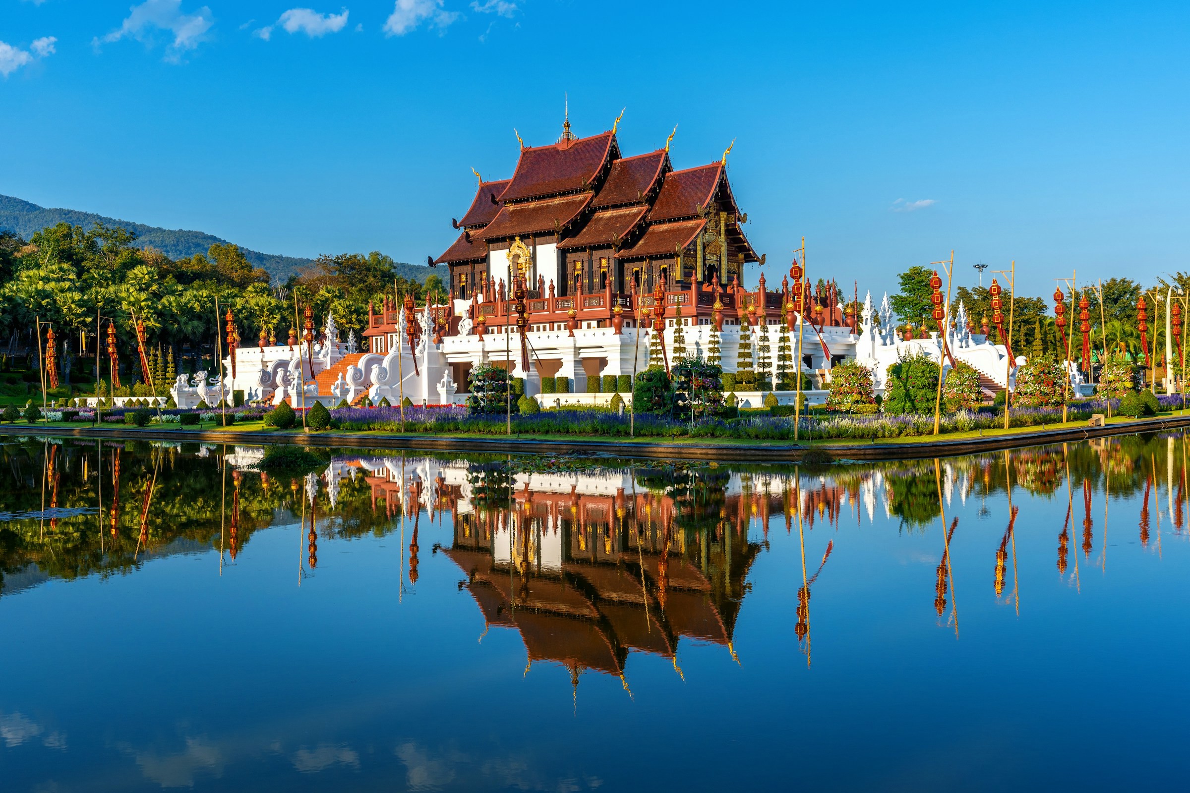 Temples in Phnom Penh with ornate wooden details are reflected in a water surface, surrounded by lush greenery and clear blue skies.