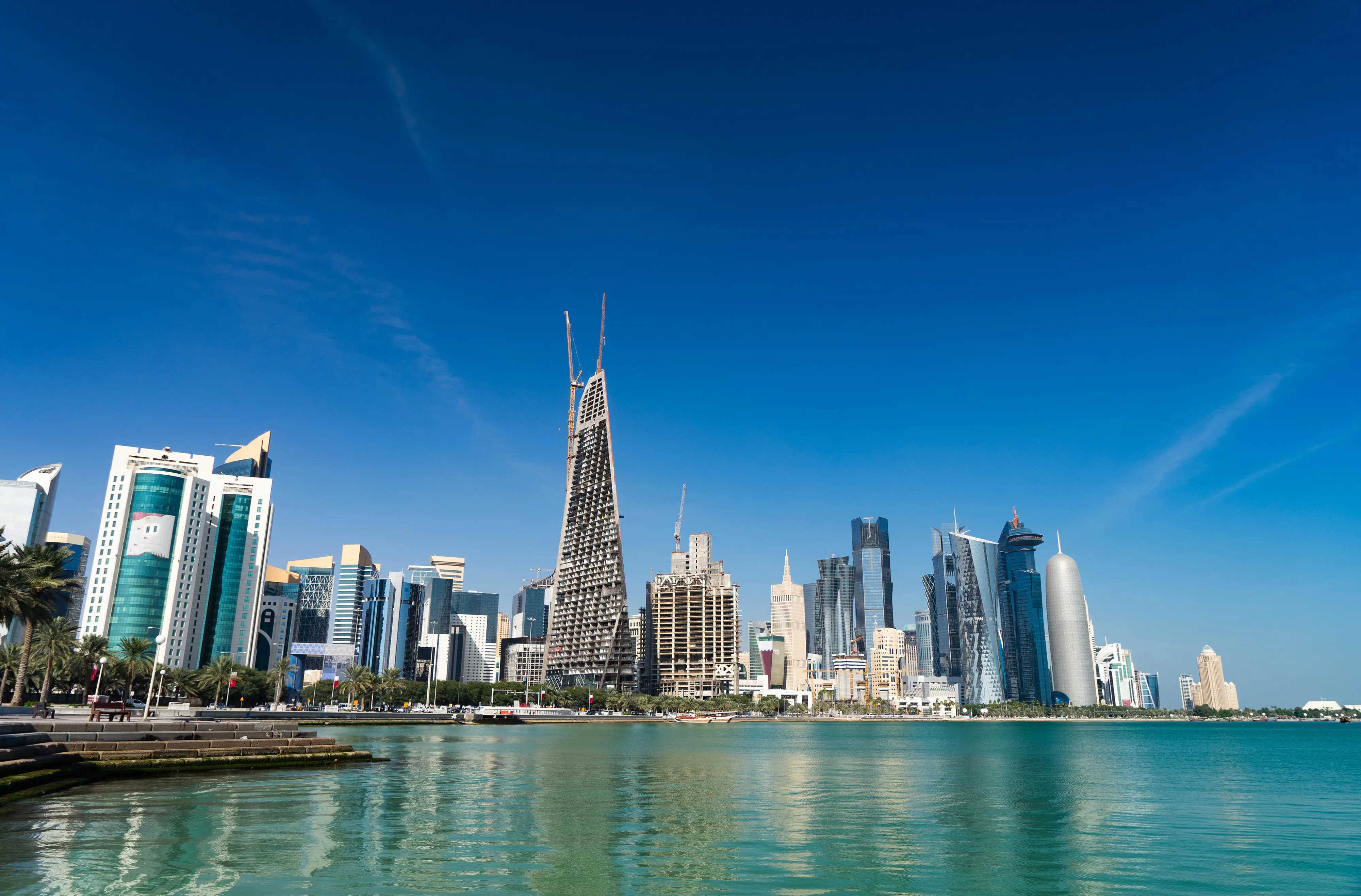 Resa till Qatar - Panoramavy över en modern stadssiluett med skyskrapor vid en klarblå himmel bredvid ett lugnt vatten.