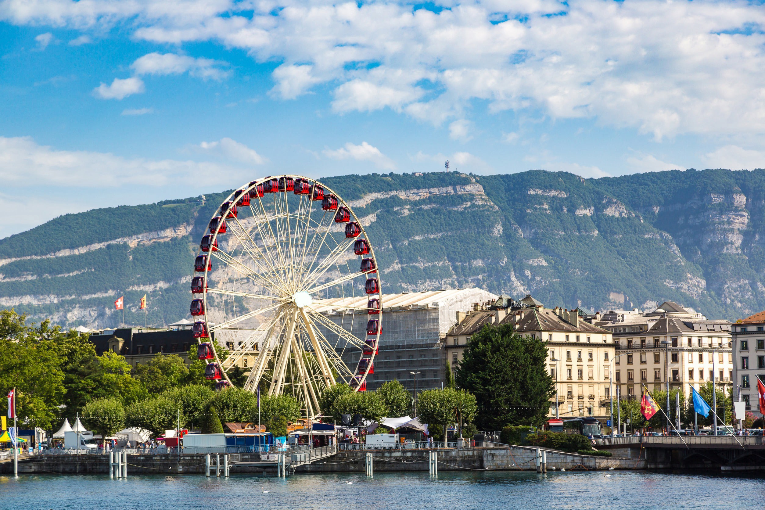 Vy över Geneve stad med byggnader och ett pariserhjul och grönska omkring sig med vackra kullar och blå himmel i bakgrunden