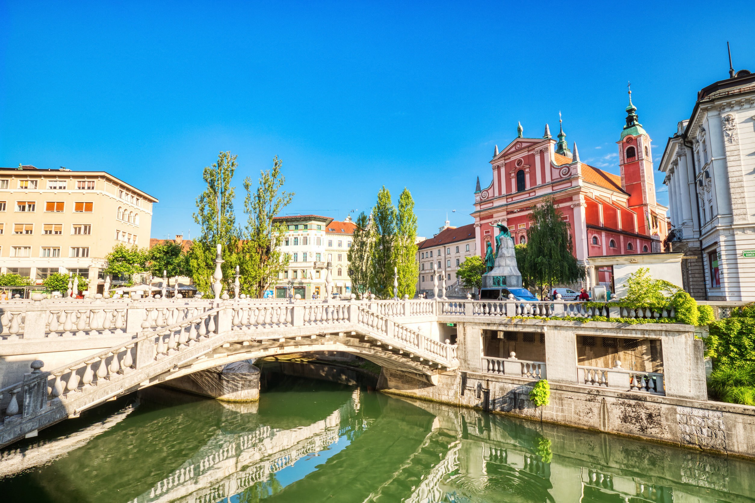 A bridge leads over a bridge towards a beautiful red church with green trees around it and a clear blue sky in the background