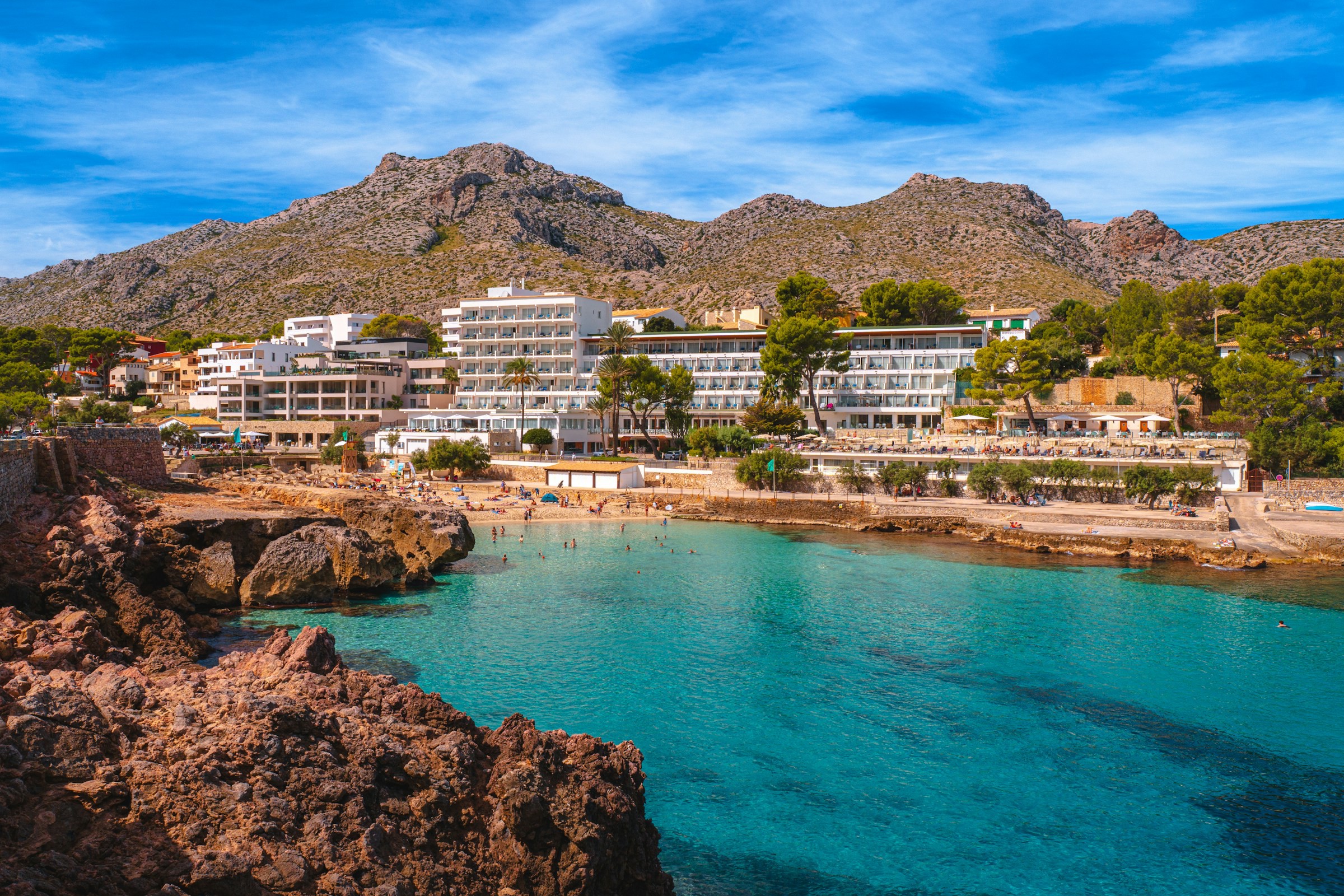 Vacker bukt med blått hav som leder upp till en strand med ett stort vitt hotell och berg på Mallorca