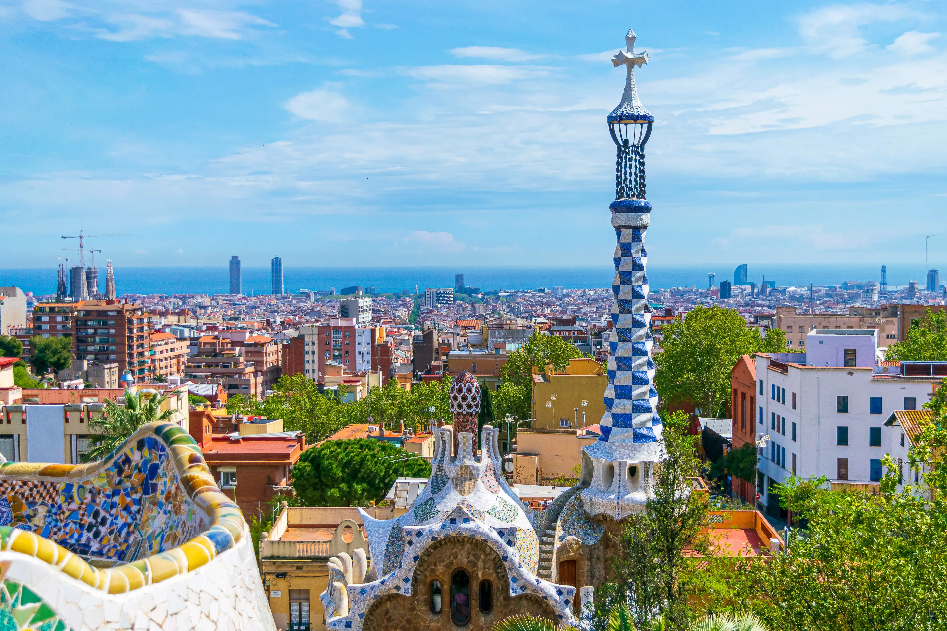 Resa till Barcelona - Utsikt över Barcelona från Park Güell med färgglada mosaiker och den ikoniska serpentinformade bänken i förgrunden, och stadens silhuett mot en klarblå himmel i bakgrunden.