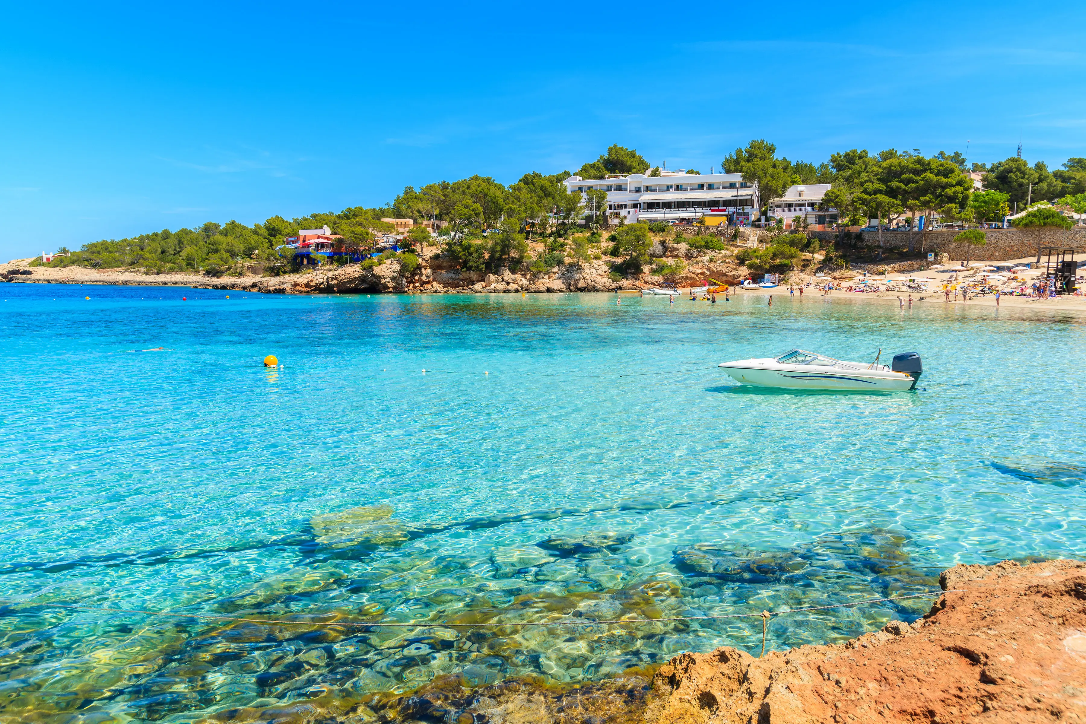 Resa till Ibiza - Klart turkos vatten vid en solig medelhavsstrand med folk, en båt och grönskande vegetation i bakgrunden.