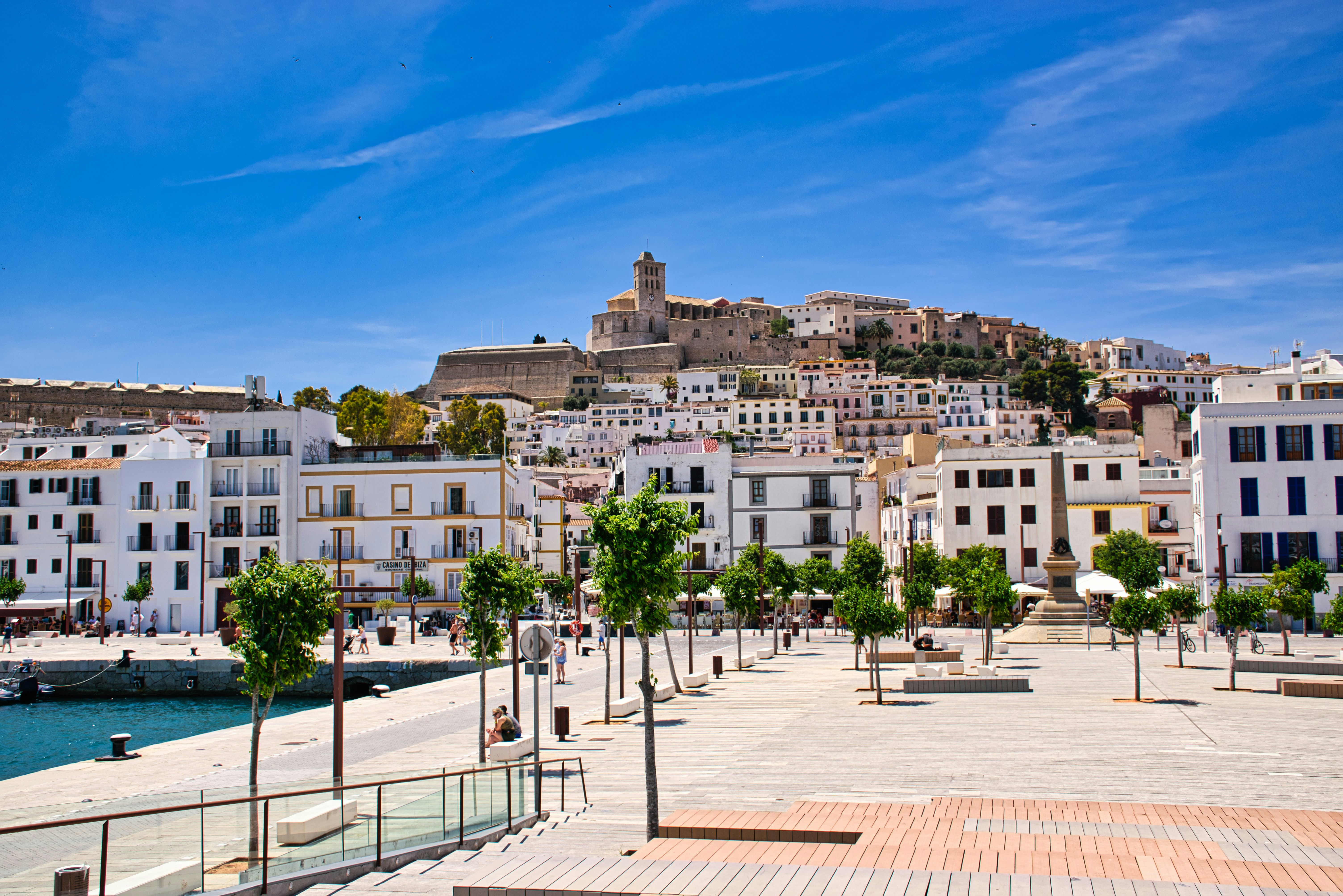 Vitkalkade byggnader och en livlig strandpromenad mot bakgrunden av en befäst stad på en kulle under en klarblå himmel på Ibiza.