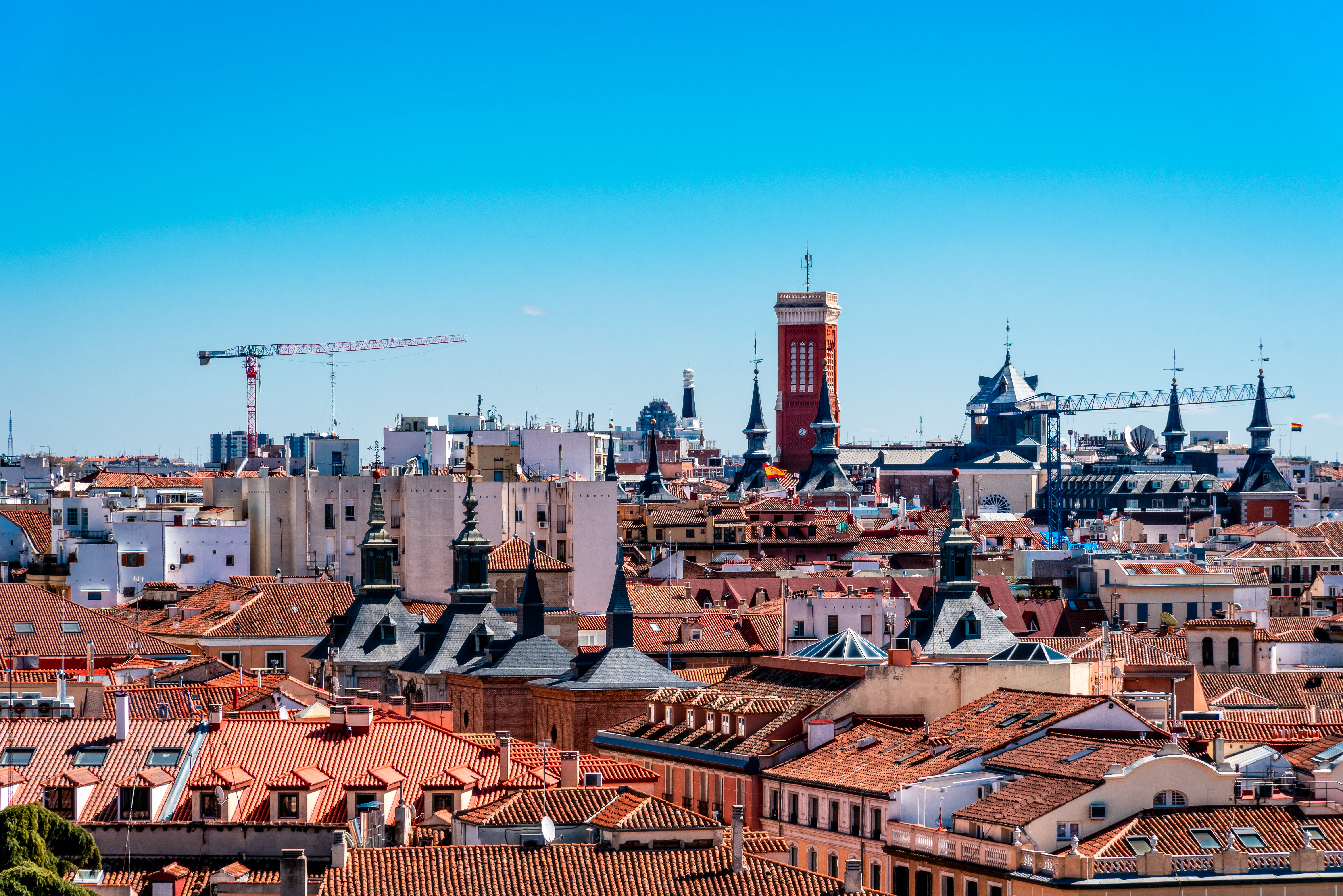 Resa till Madrid - Panoramavy över en livlig stad med röda tak, byggkranar och moderna byggnader mot en klarblå himmel, som illustrerar en blandning av historisk och modern arkitektur.