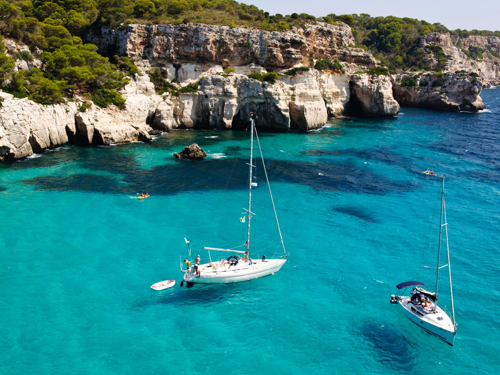 Resa till Menorica - Segelbåtar på turkosblått vatten vid klippiga kusten med frodig grönska.