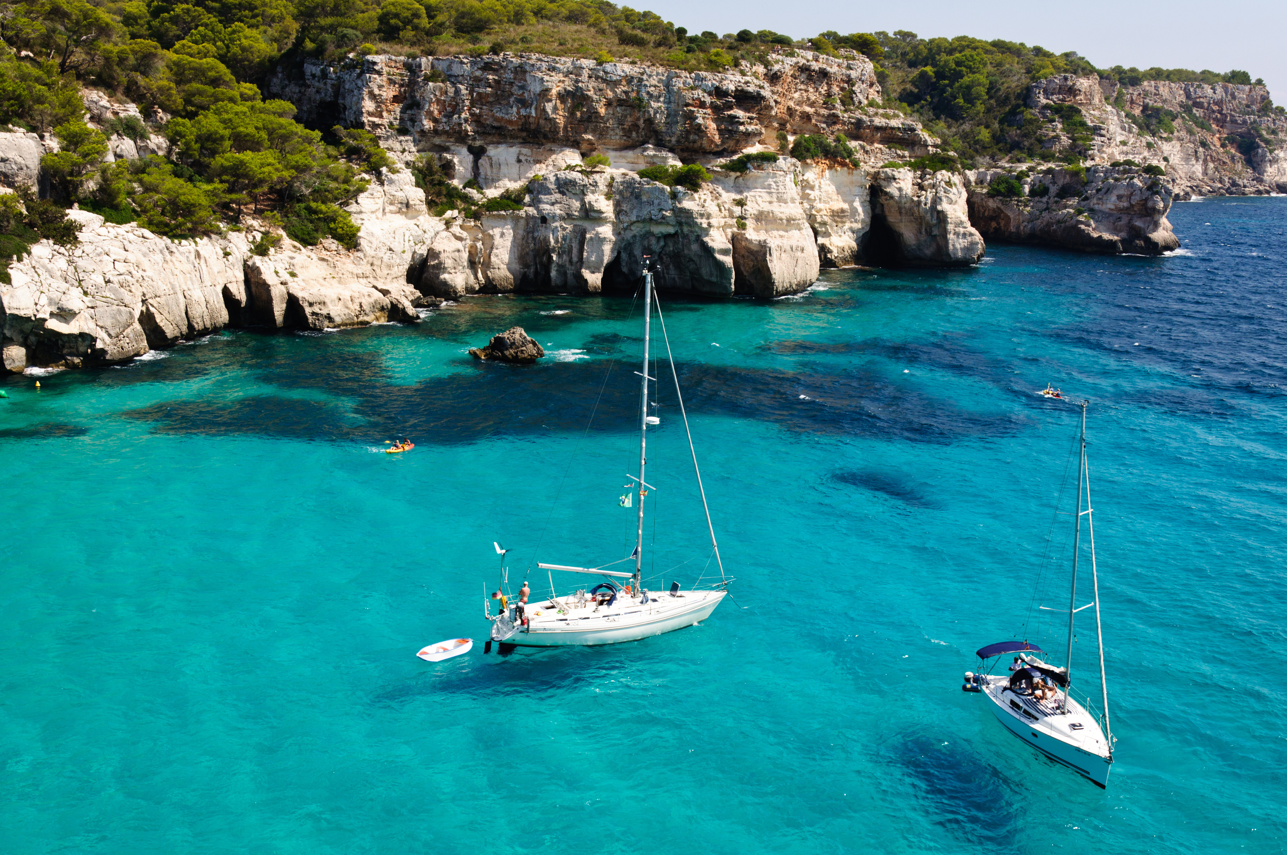 Resa till Menorica - Segelbåtar på turkosblått vatten vid klippiga kusten med frodig grönska.