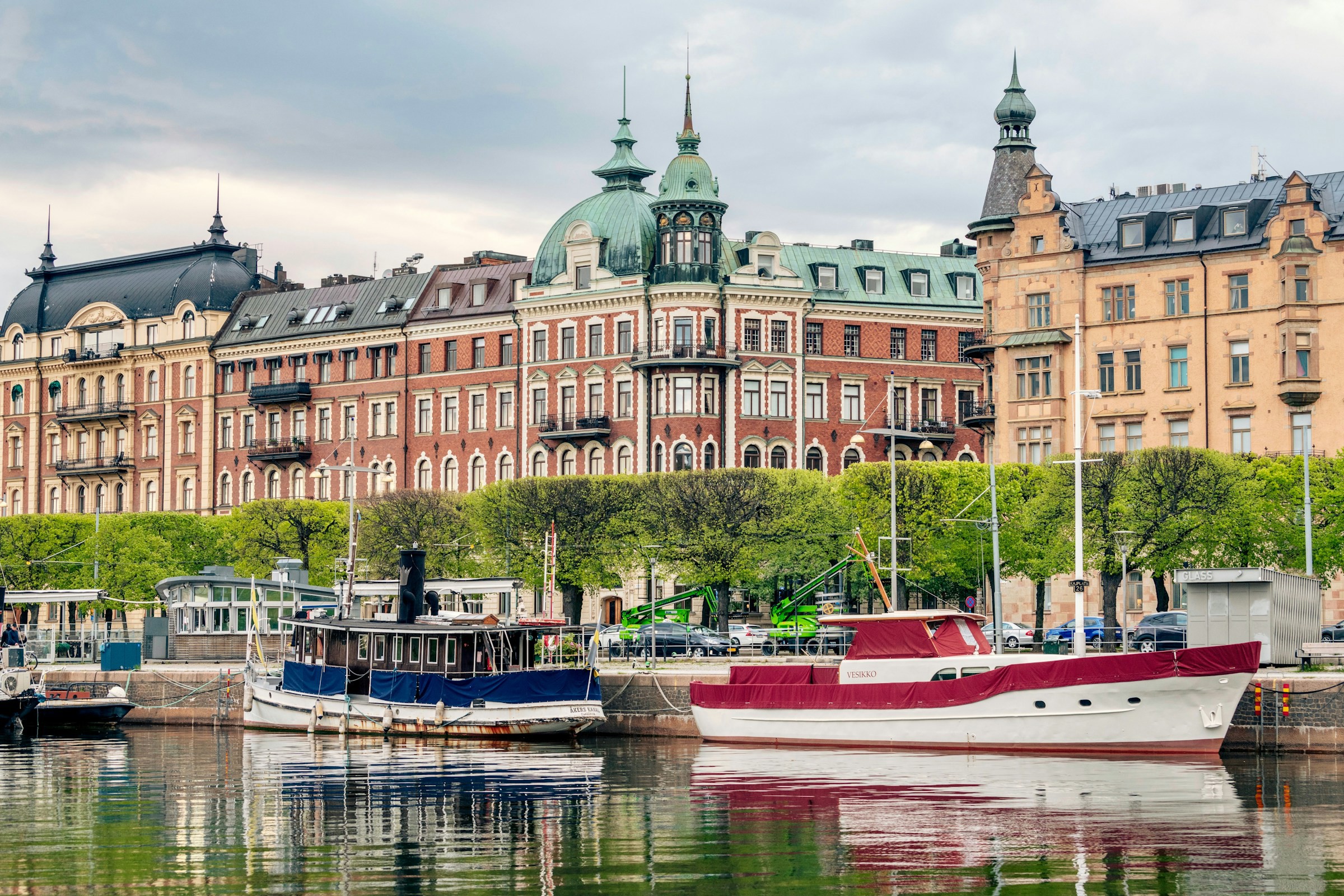 Klassiska byggnader och fritidsbåtar vid vattnet i Stockholm, Sverige, spegling av båtar i lugnt vatten med grönskande träd i förgrunden.