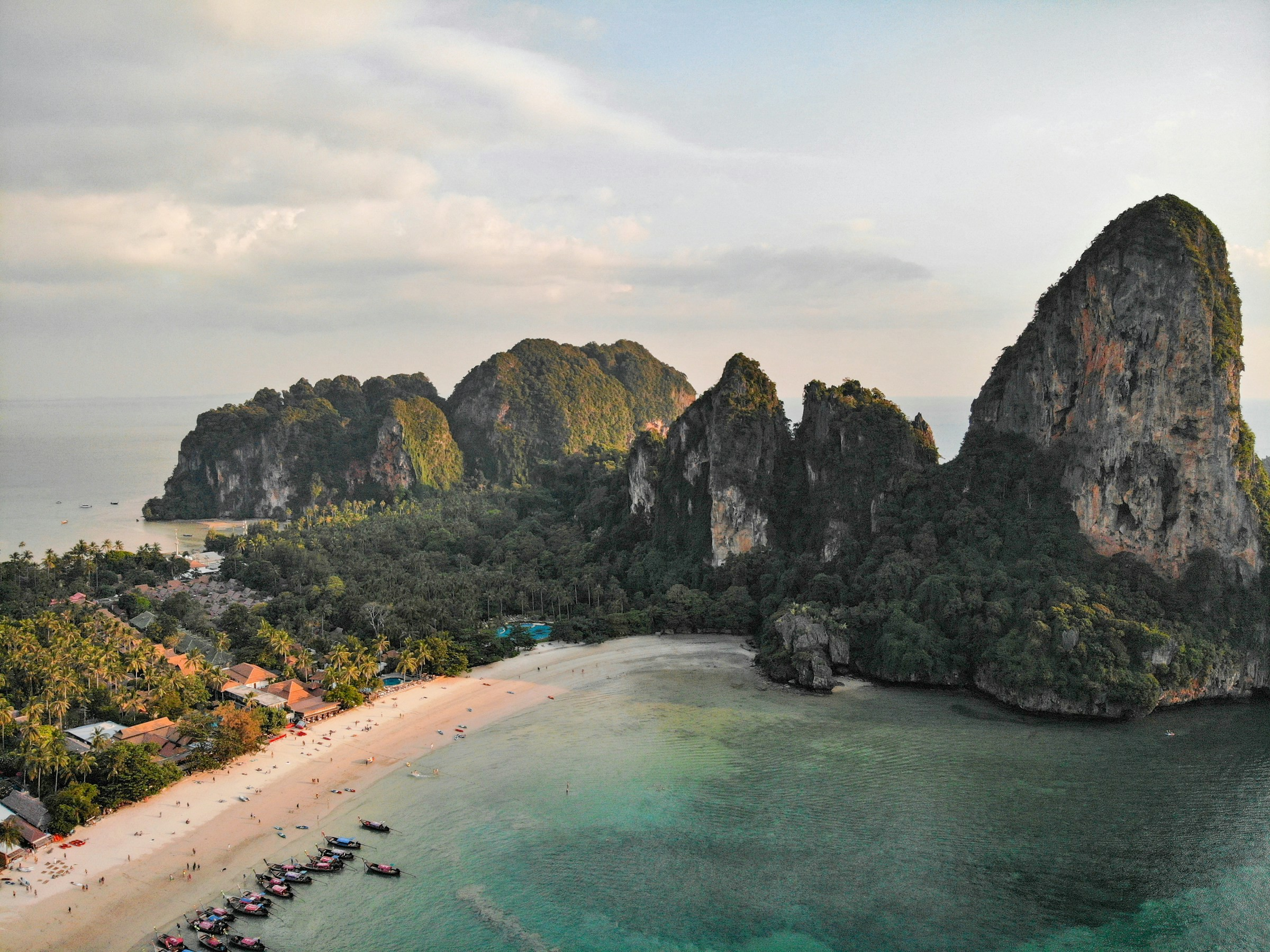 Fågelvy över bukt med strand och vackert hav som möter höga berg med grönska och blå himmel i bakgrunden i Krabi