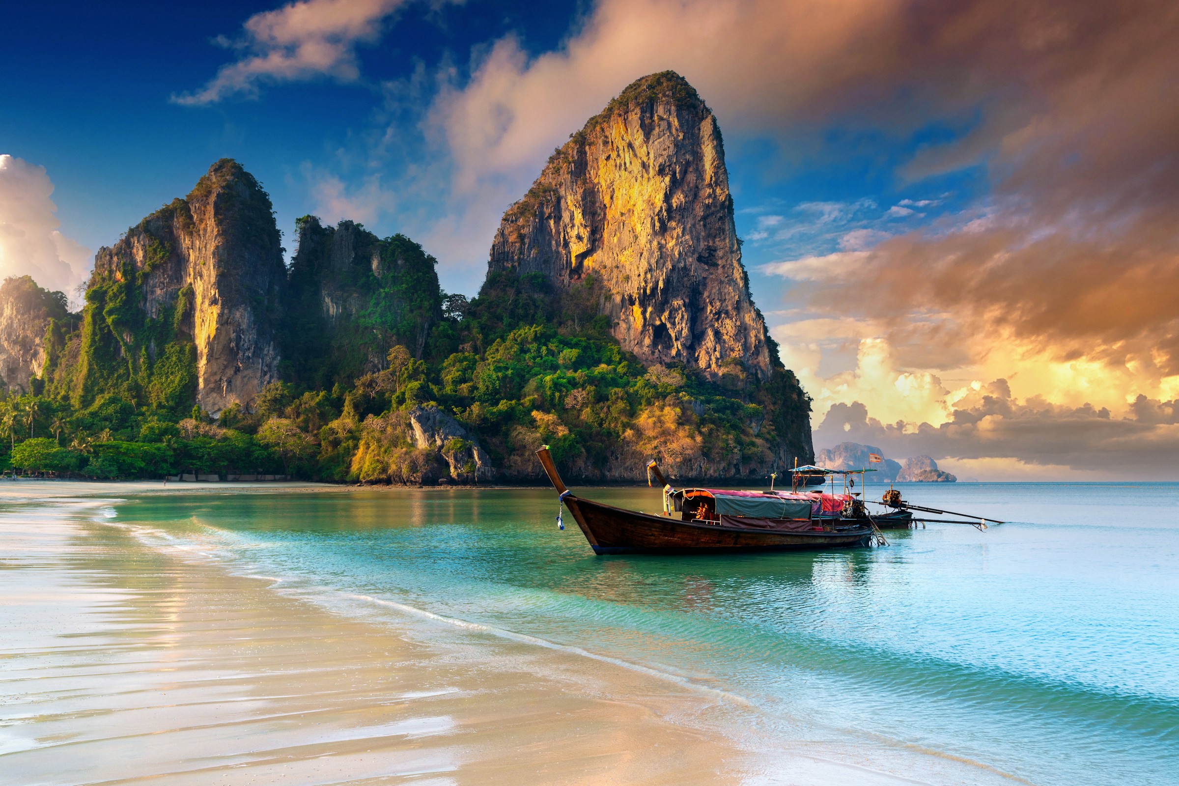 Tidig skymning vid en strand som möter blått hav och en longtail-båt med berg i bakgrunden i Thailand