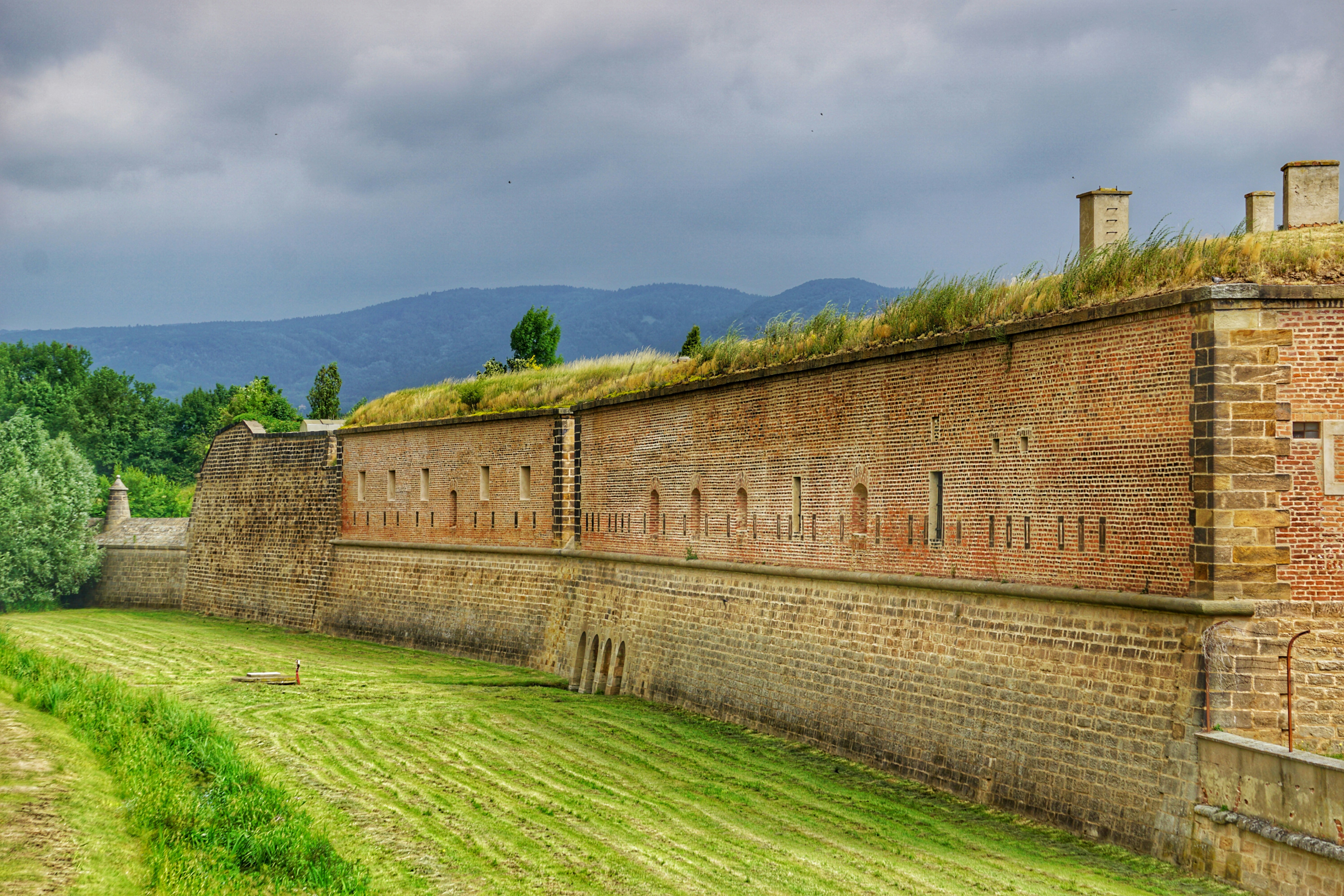 Gammal tegelfästning med grästak och öppet grönt fält framför under molnig himmel, historiskt försvarsverk i Tjeckien.