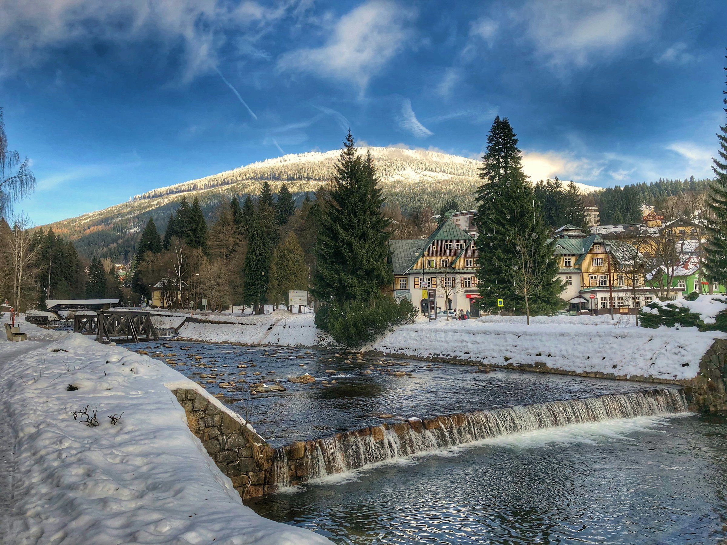 En flod går igenom en liten snötäckt by i Spindleruv Mlyn med berg och blå himmel i bakgrunden