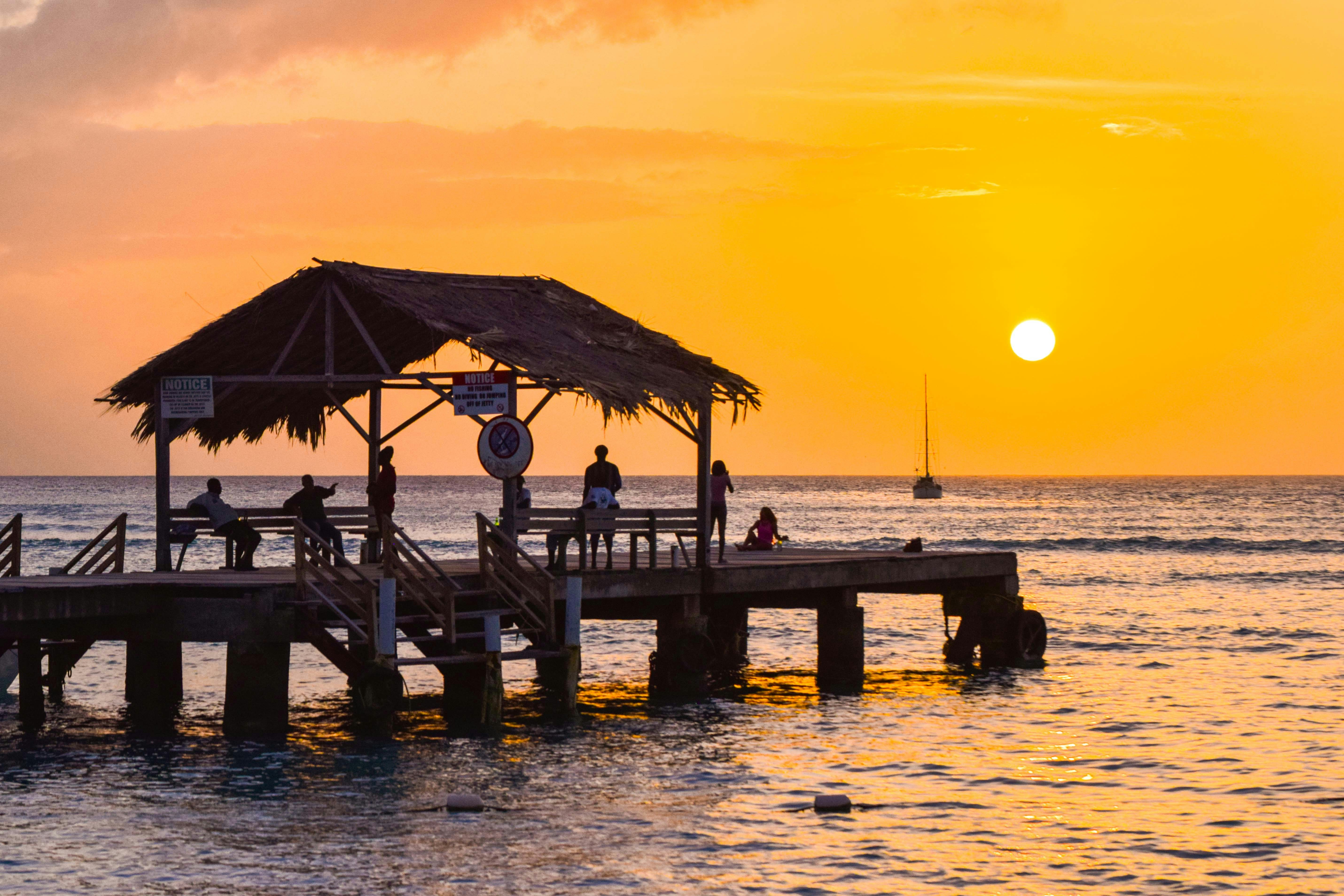 Solnedgång vid havet med människor på en träbrygga med ett stråtak, och en segelbåt i fjärran i Trinidad, Tobago.